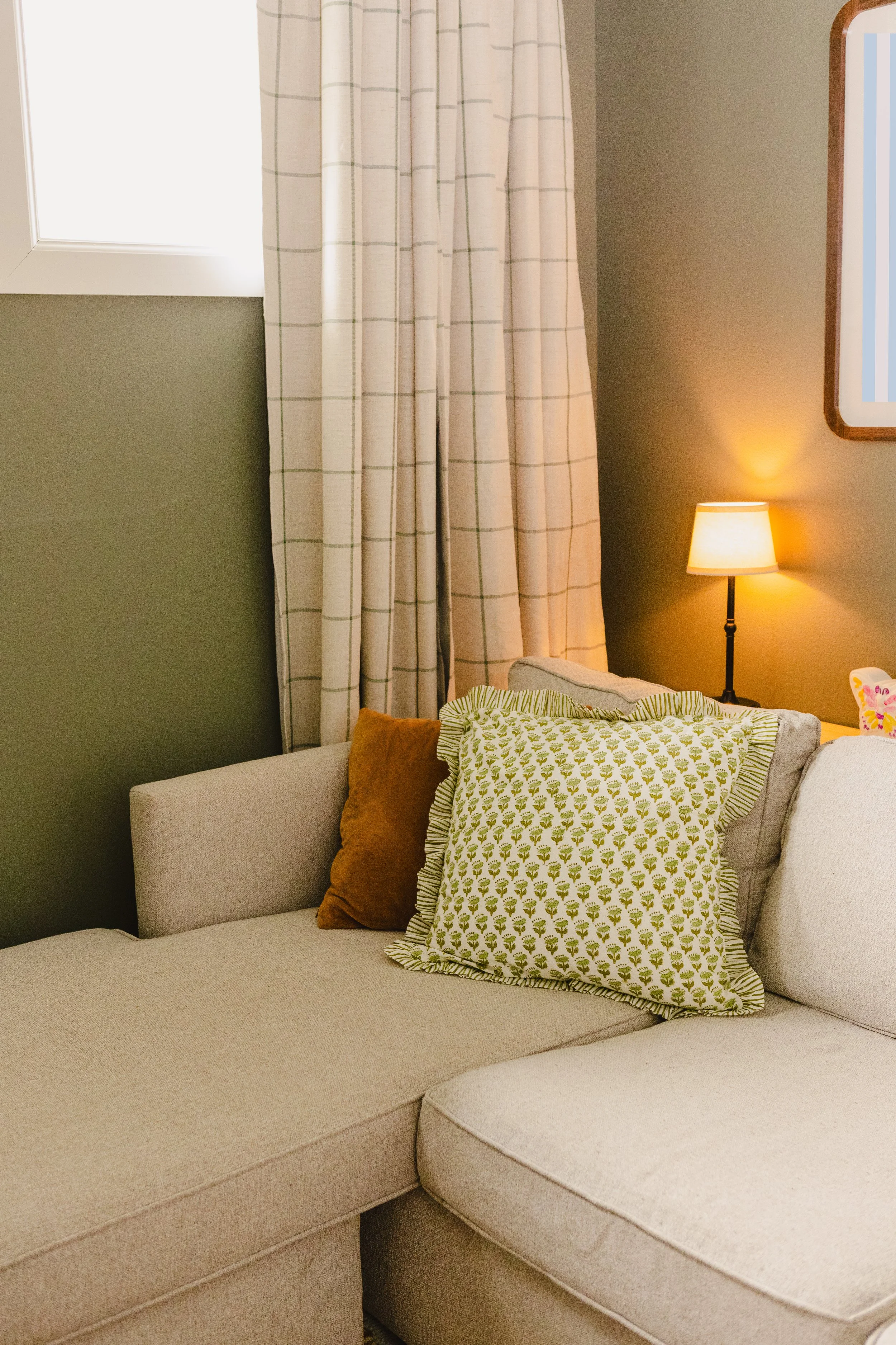 A living room with a beige sectional sofa and cushions, a side table with a lamp, a window with curtains, and a framed mirror on the wall.