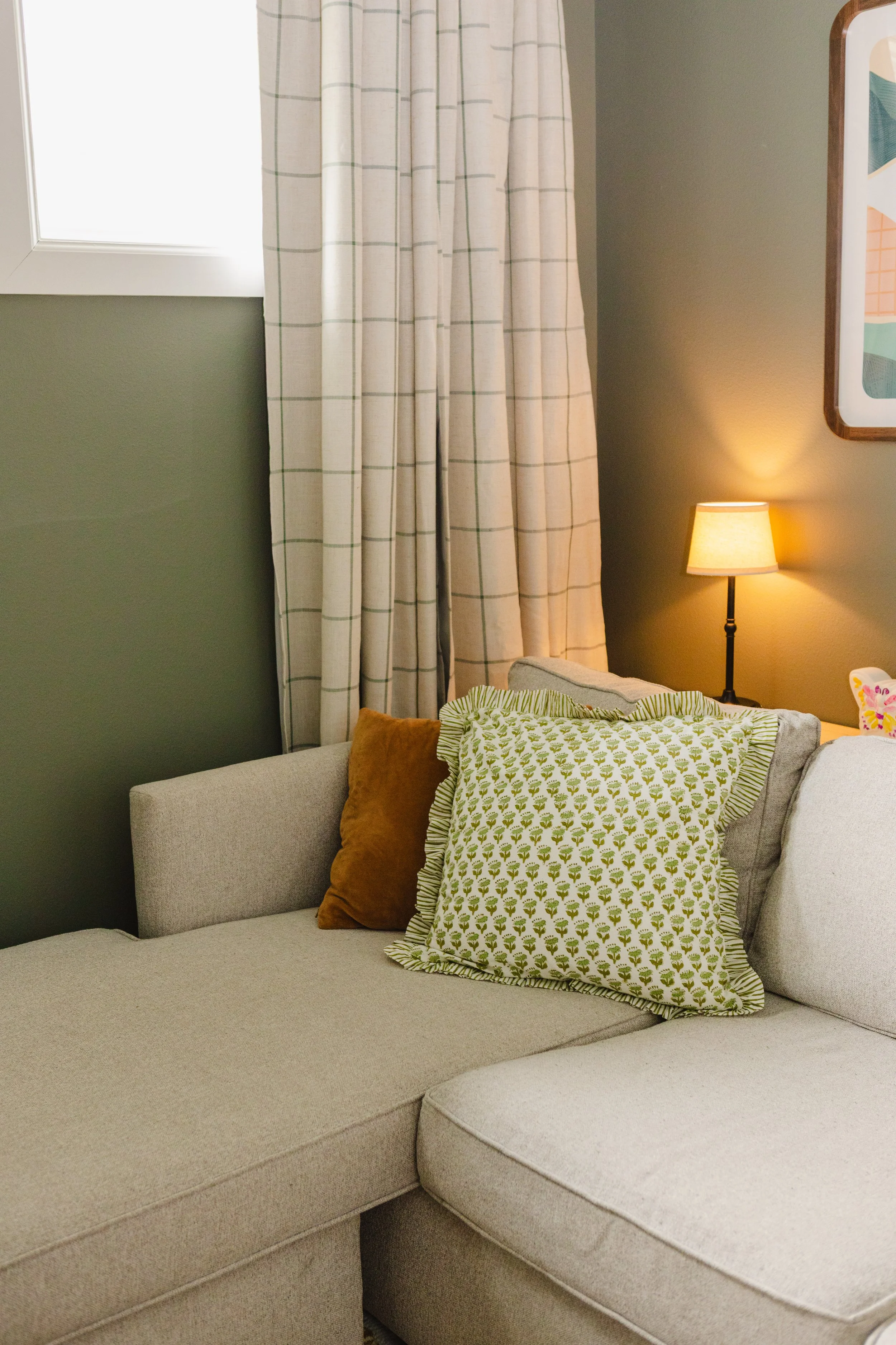 A beige sofa with decorative pillows, a green-and-white patterned pillow, a brown pillow, and a beige pillow, alongside a lamp on a side table.