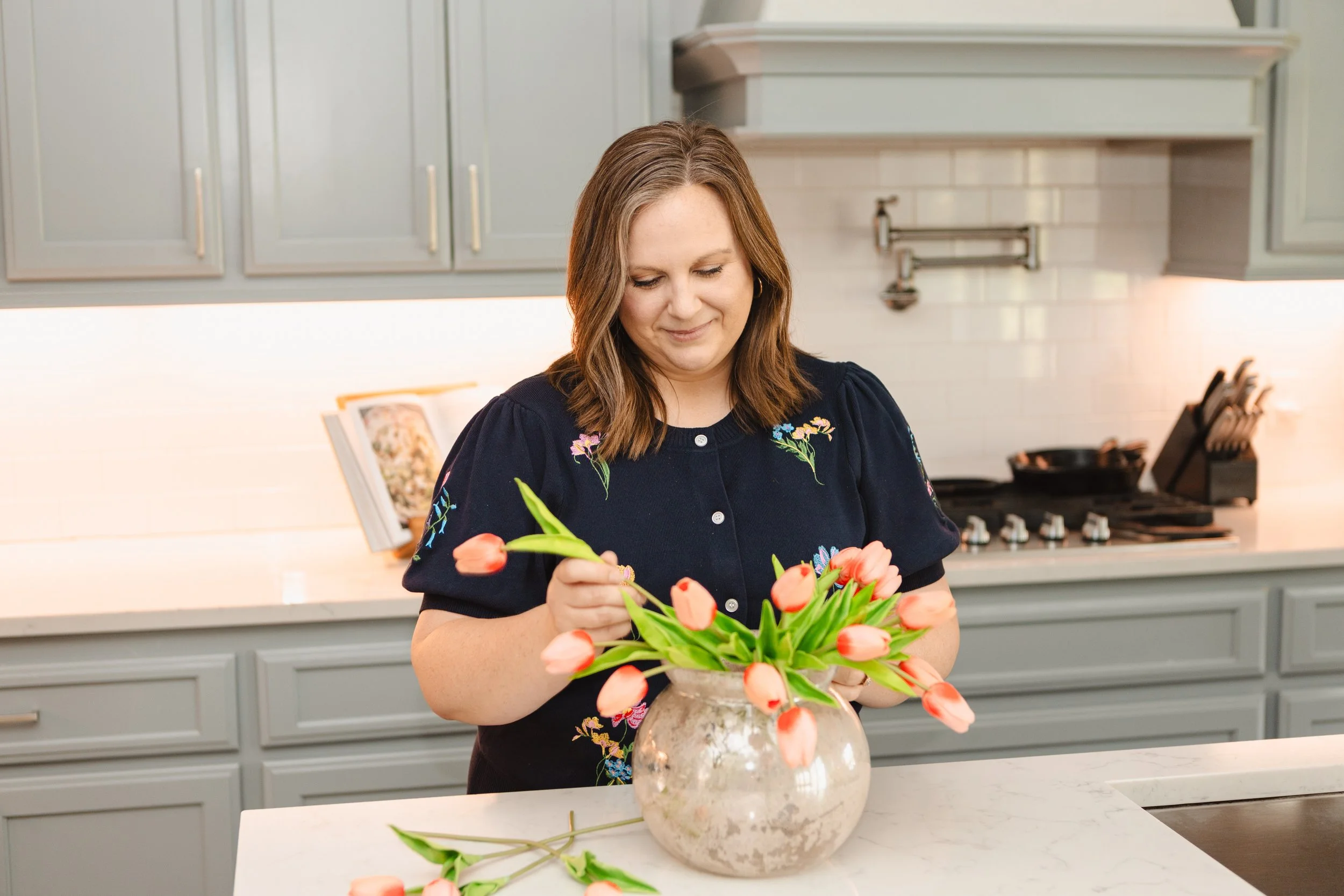 A woman arranging a bouquet of orange tulips in a marble vase in a modern kitchen.