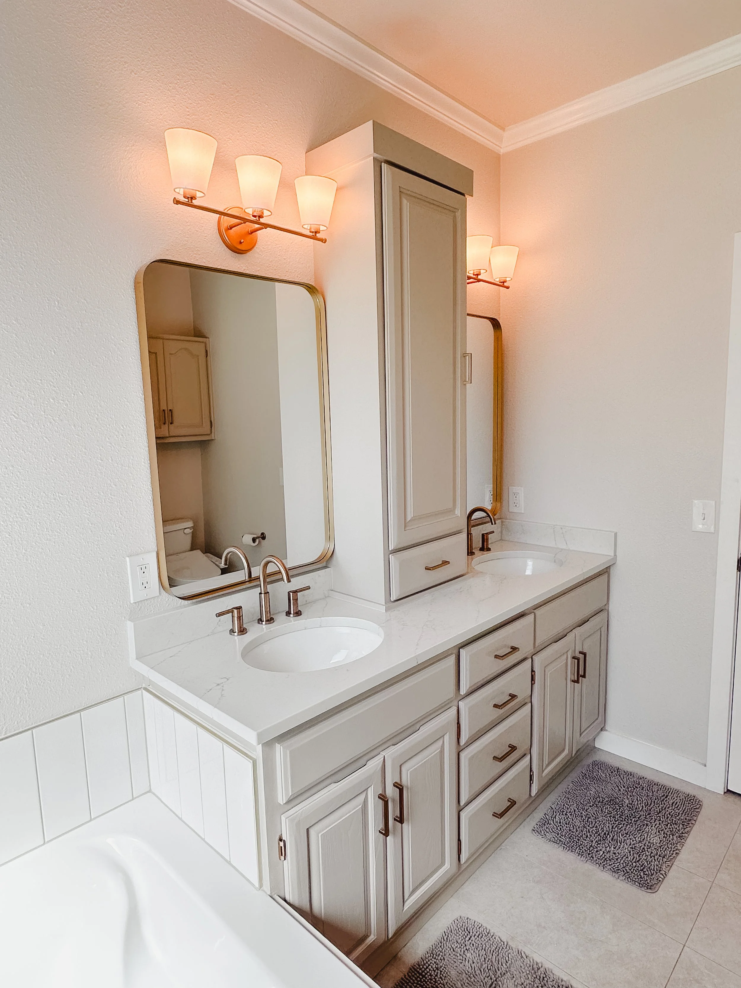 Bathroom vanity with two sinks, marble countertop, upper cabinets, a large mirror, wall-mounted light fixtures with four bulbs, and bathroom mats on the floor.