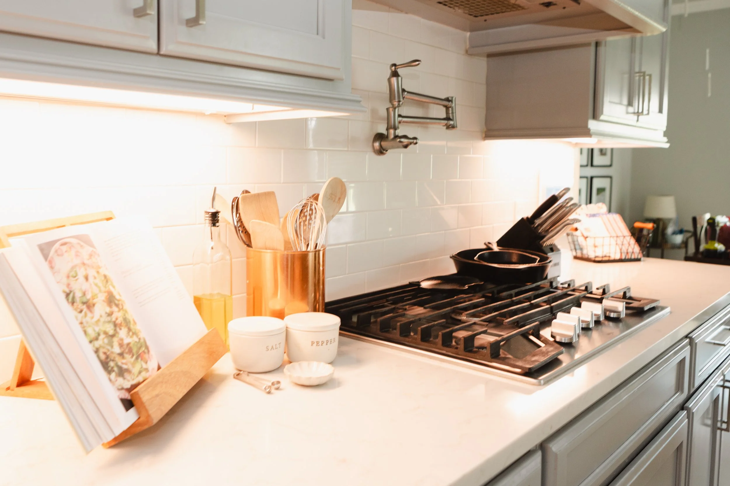 A kitchen countertop with various cooking utensils, open cookbook, salt and pepper containers, and a stove with cast iron pans.