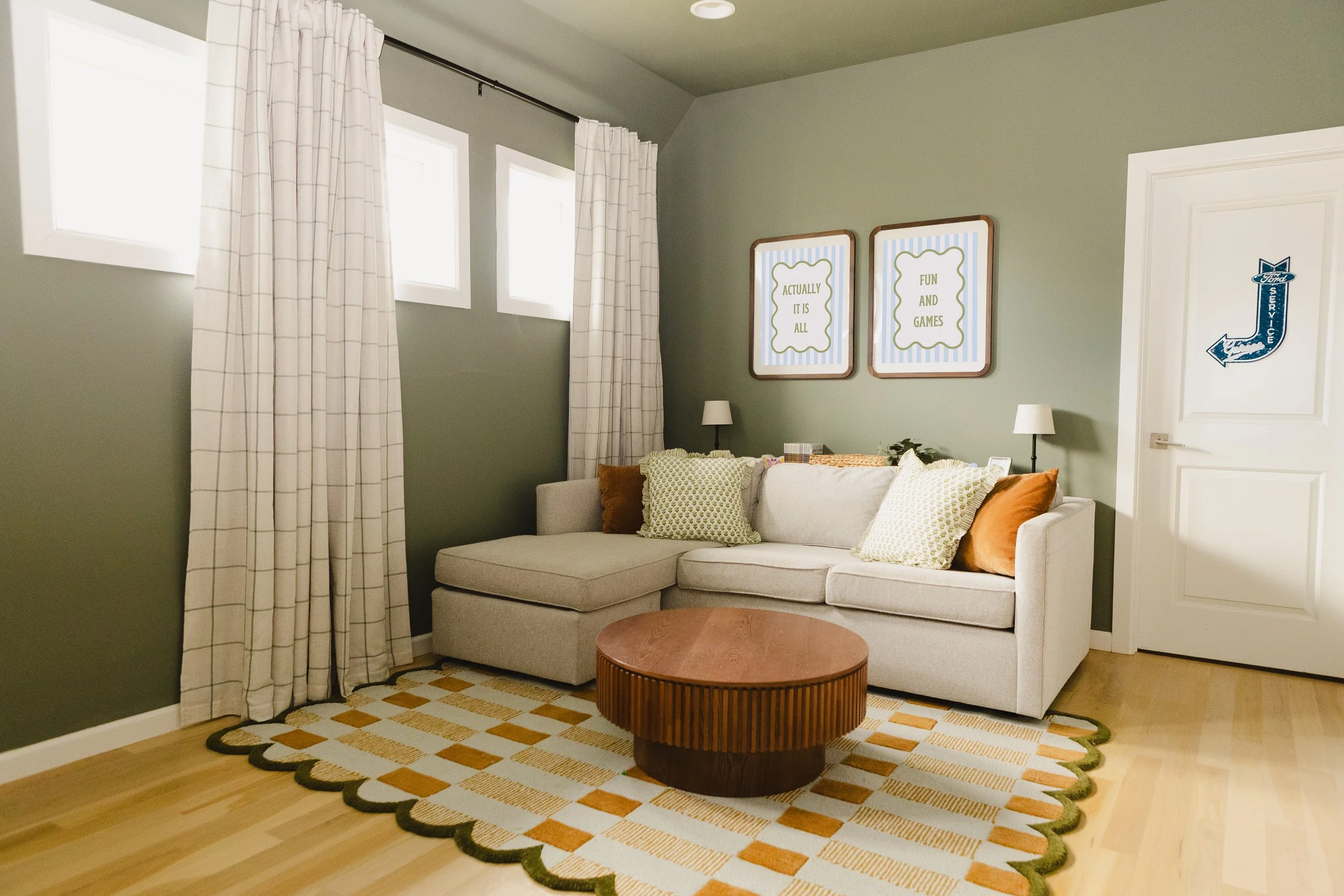 Living room with green walls, white curtains, a beige sectional sofa with patterned pillows, a round wooden coffee table, a yellow and orange checkered rug, two framed quotes on the wall, and a door with a blue arrow sign.