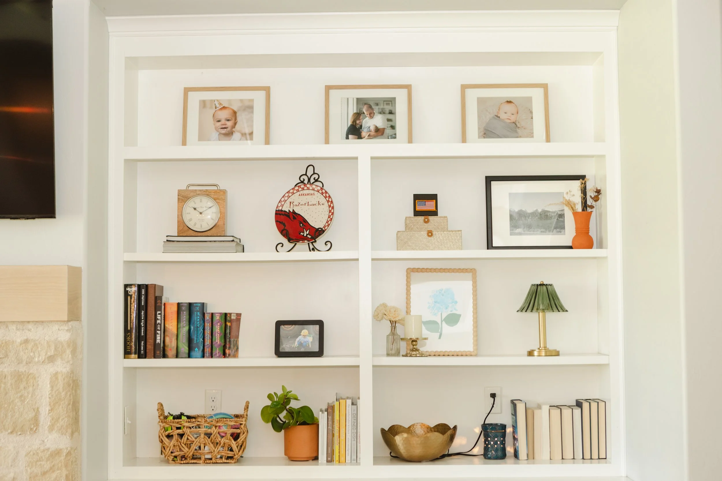 White bookshelf with framed family photos on top, a clock, a Nazarene emblem, framed artwork, a green lamp, books, plants, and decorative bowls and baskets.