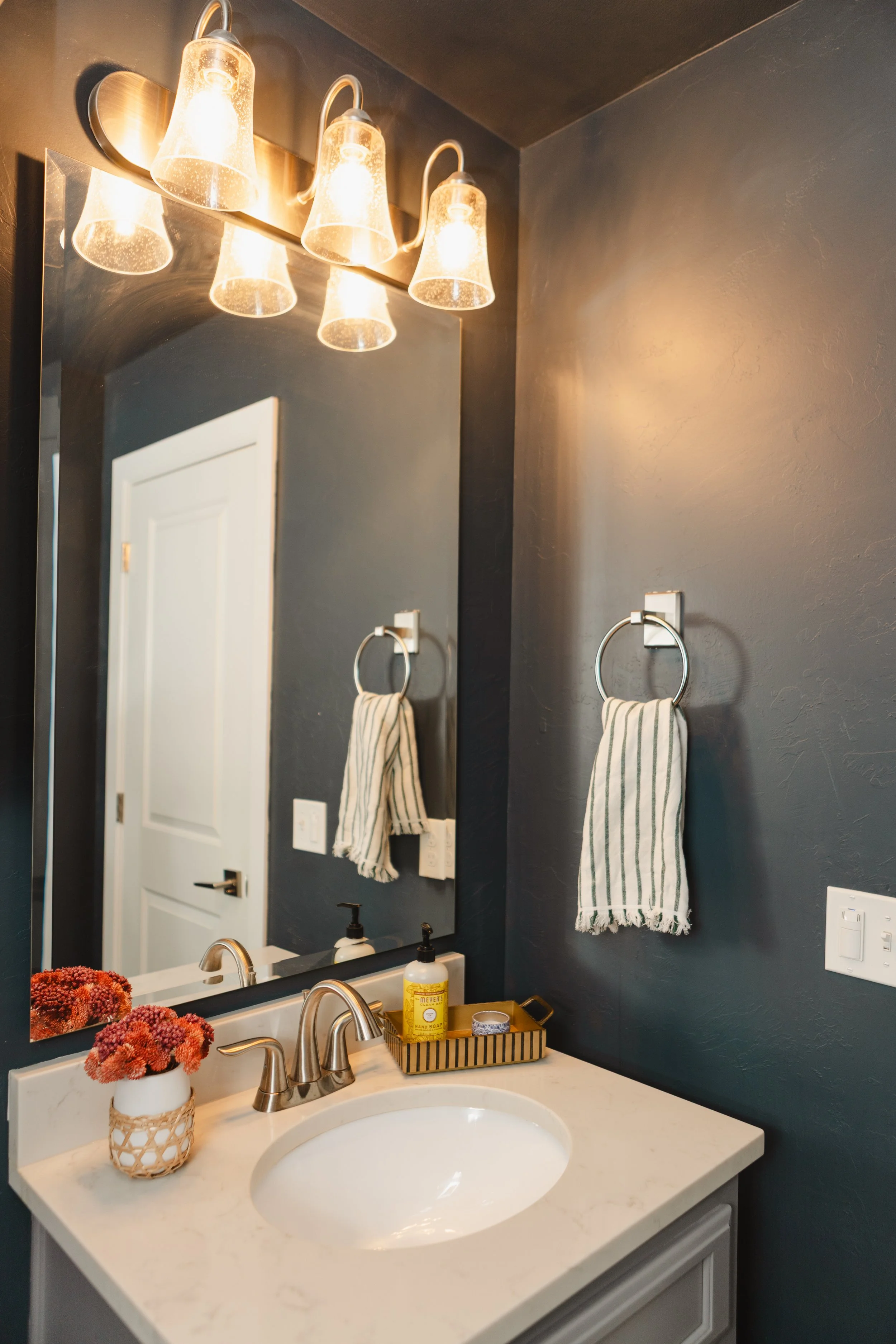 Bathroom vanity with a white countertop, a sink, a mirror, and a wall-mounted light fixture with four glass shades. A vase with pink flowers, a soap dispenser, and a tray with toiletries sit on the counter. Towel hooks with striped towels are on the wall beside the mirror.