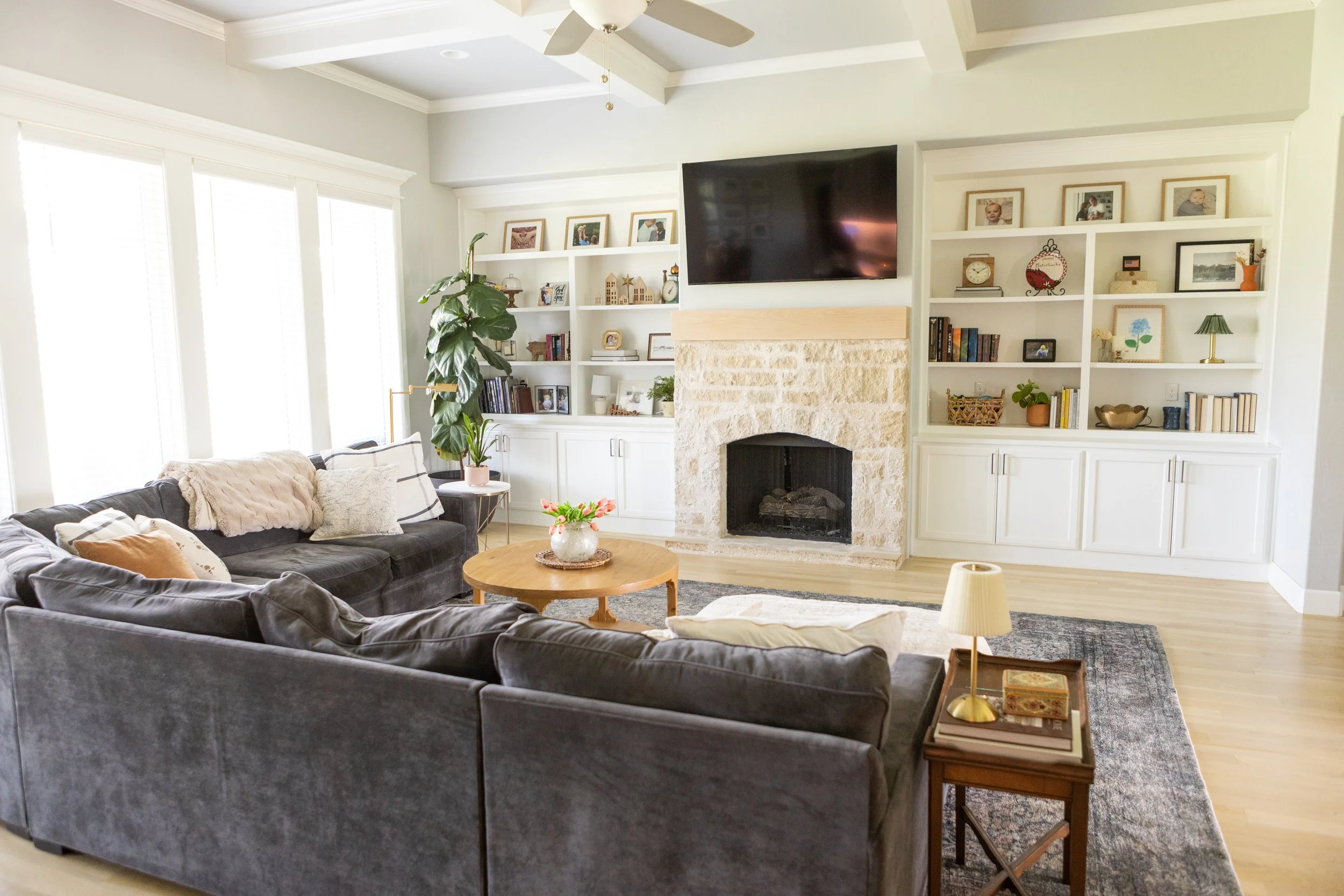 A living room with a dark gray sectional sofa adorned with patterned and solid throw pillows, a round wooden coffee table with a flower vase, a white fireplace with a large flat-screen TV mounted above, built-in white bookshelves filled with framed photos, books, and decorative items, a large leafy plant near the window, and a beige rug.