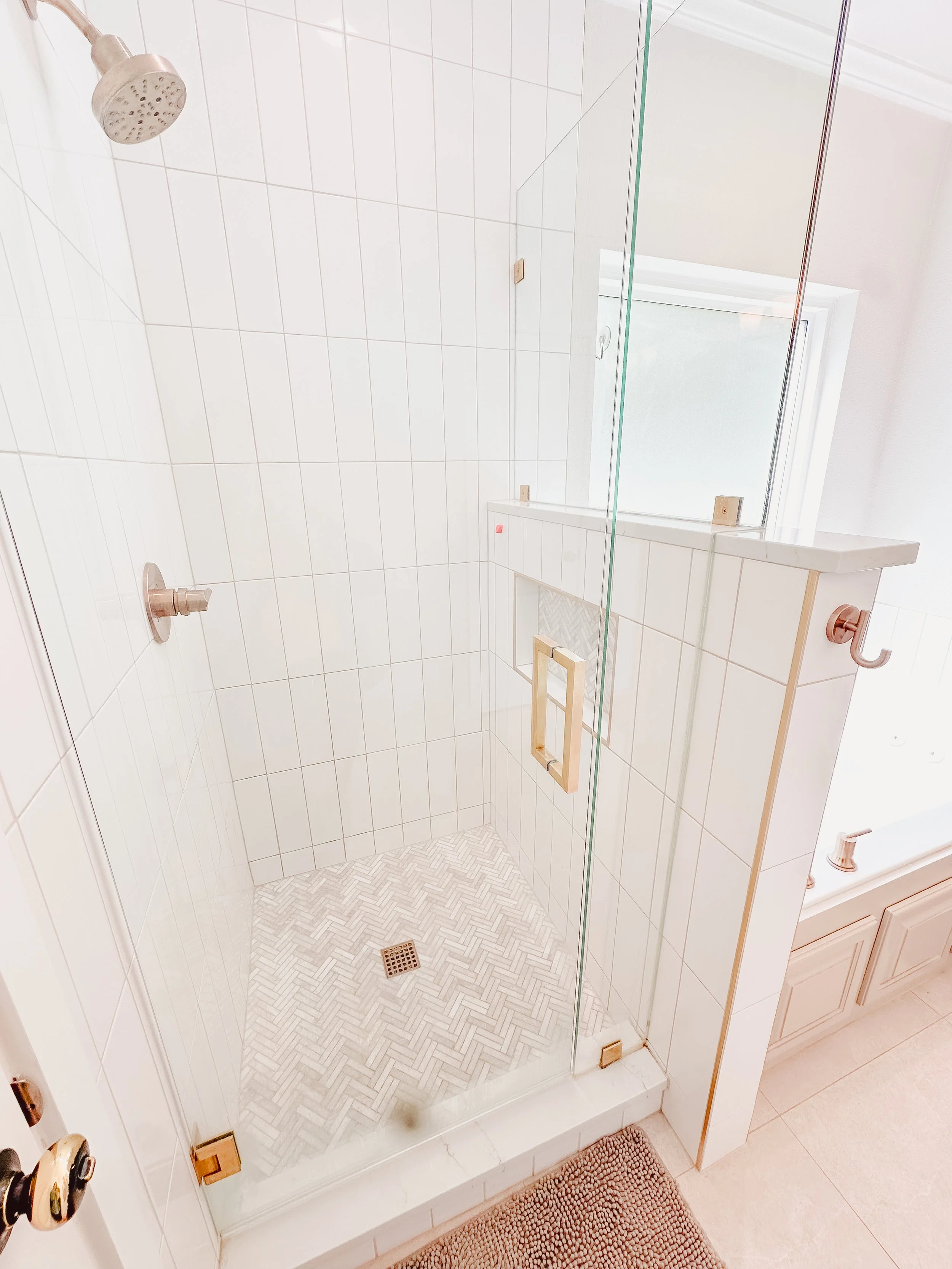 A small bathroom shower with white tiled walls, a showerhead, and a glass door with a wooden handle, situated next to a window with white trim. The shower floor has a herringbone tile pattern with a drain in the center, and there's a beige rug outsid