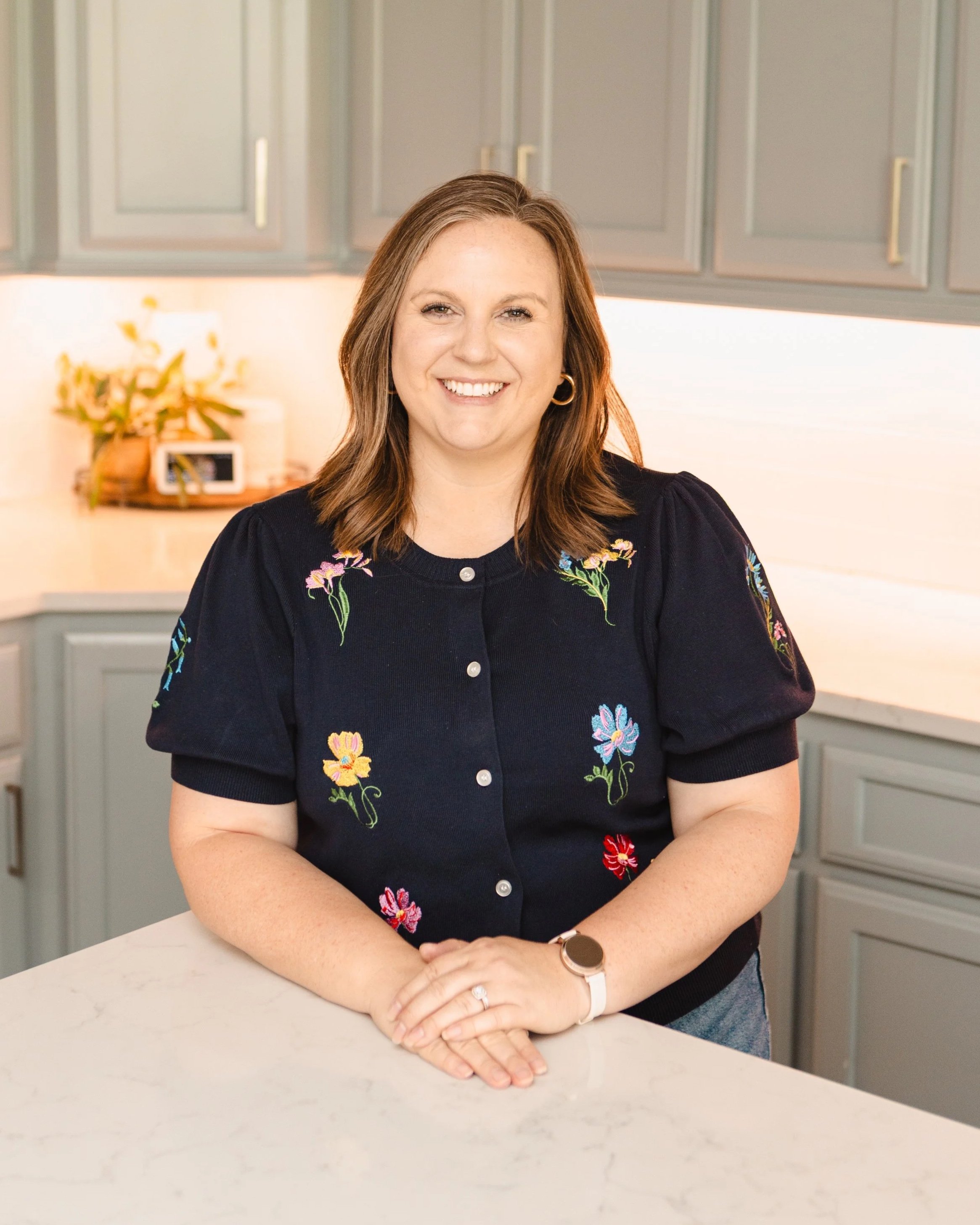 A woman with brown hair smiling, sitting at a light-colored countertop in a kitchen with gray cabinets, wearing a black embroidered top and a wristwatch.