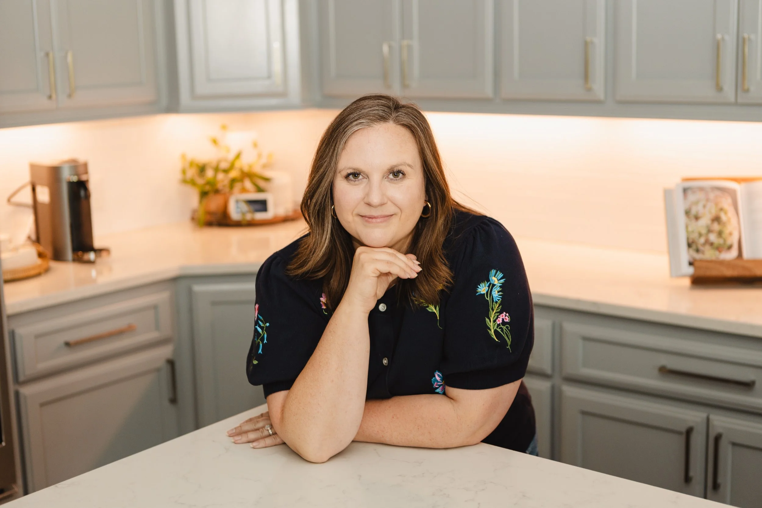 A woman with long brown hair, wearing a black top with floral embroidery, is sitting at a kitchen counter with her chin resting on her hand, smiling softly at the camera.