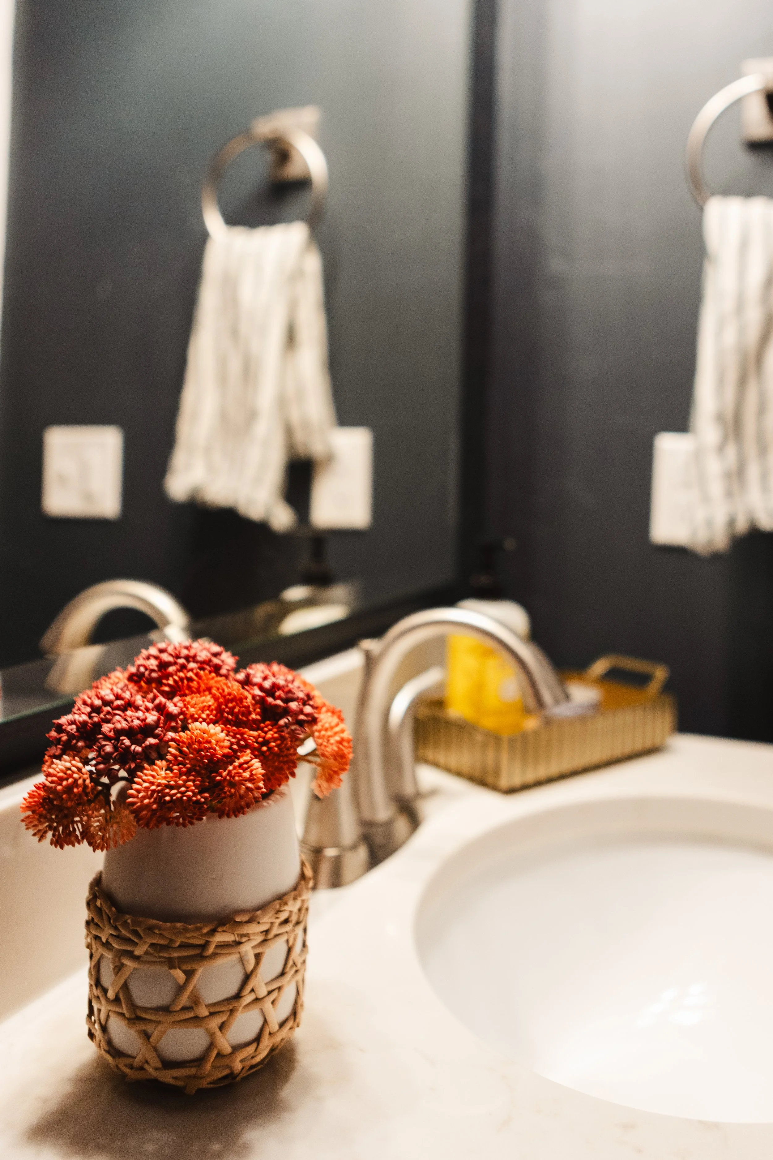 A white ceramic vase with a woven cover holding red and orange flowers on a bathroom sink counter in front of a black wall with towel hooks and a towel.