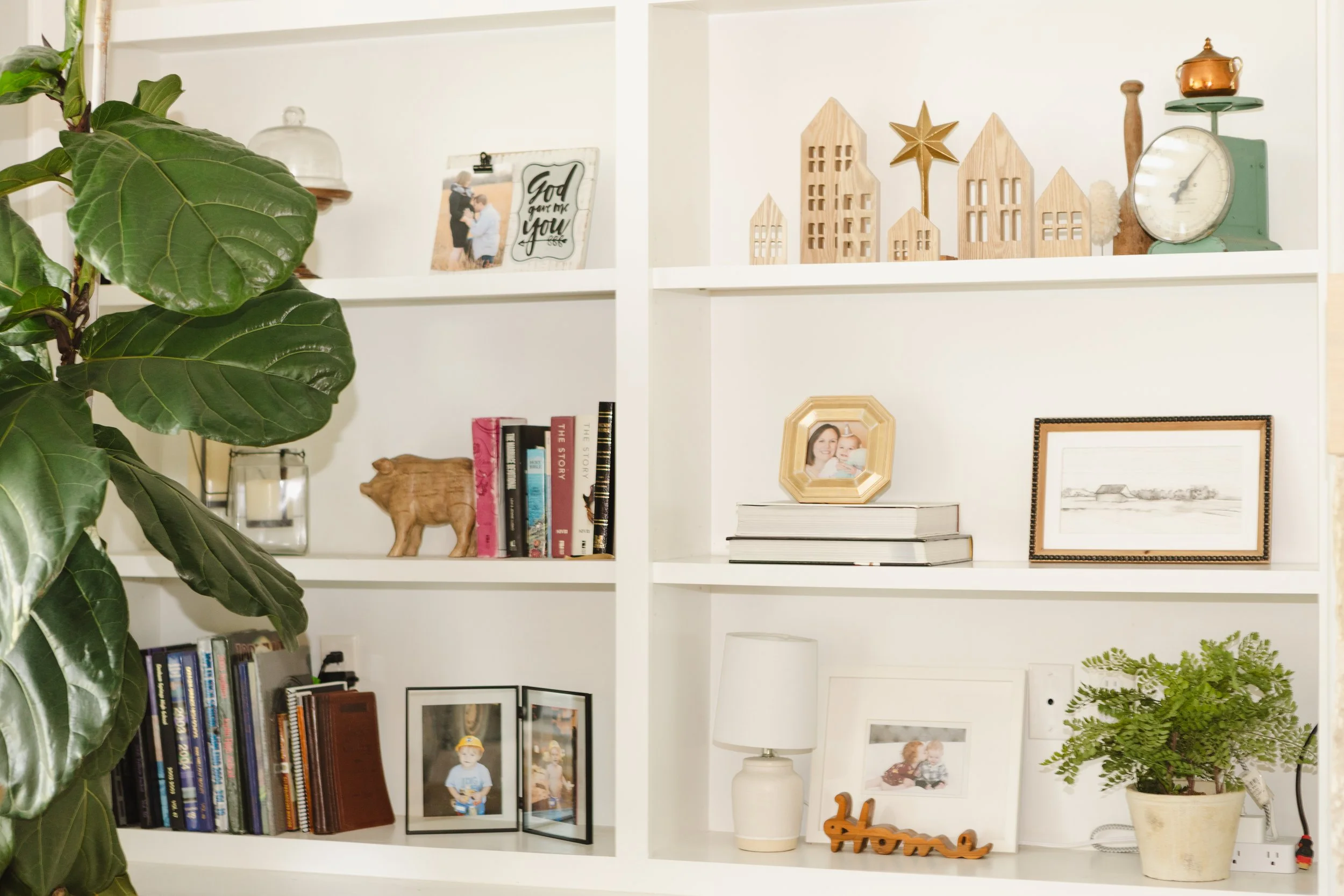 White bookshelf with decorative items, framed photographs, potted plant, books, and a small wooden 'home' sign.