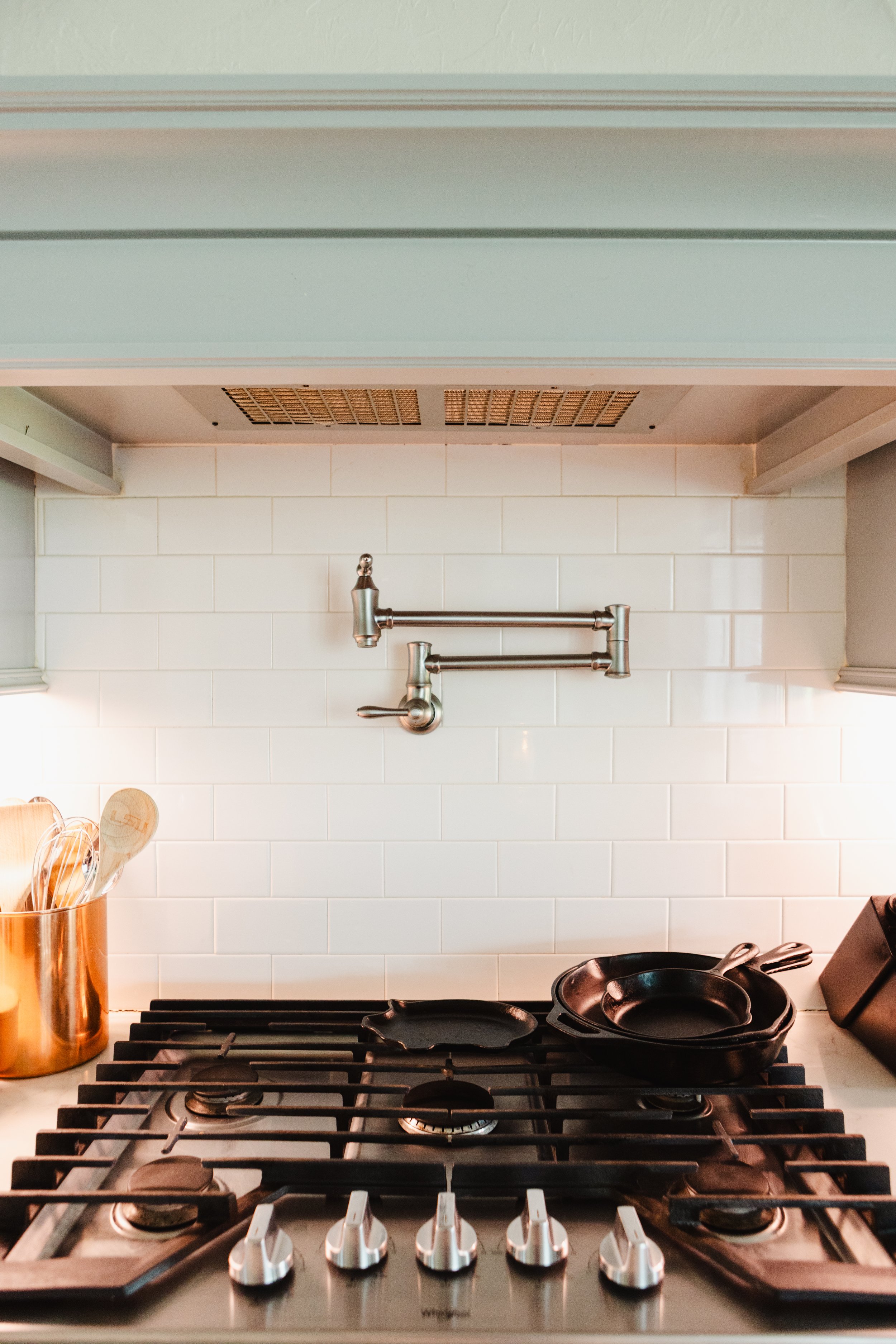 View of a modern kitchen stovetop with four burners and a black cast iron skillet on the right burner. Behind the stovetop is a white subway tile backsplash, with a metallic kitchen faucet mounted above. To the left, a copper utensil holder contains 