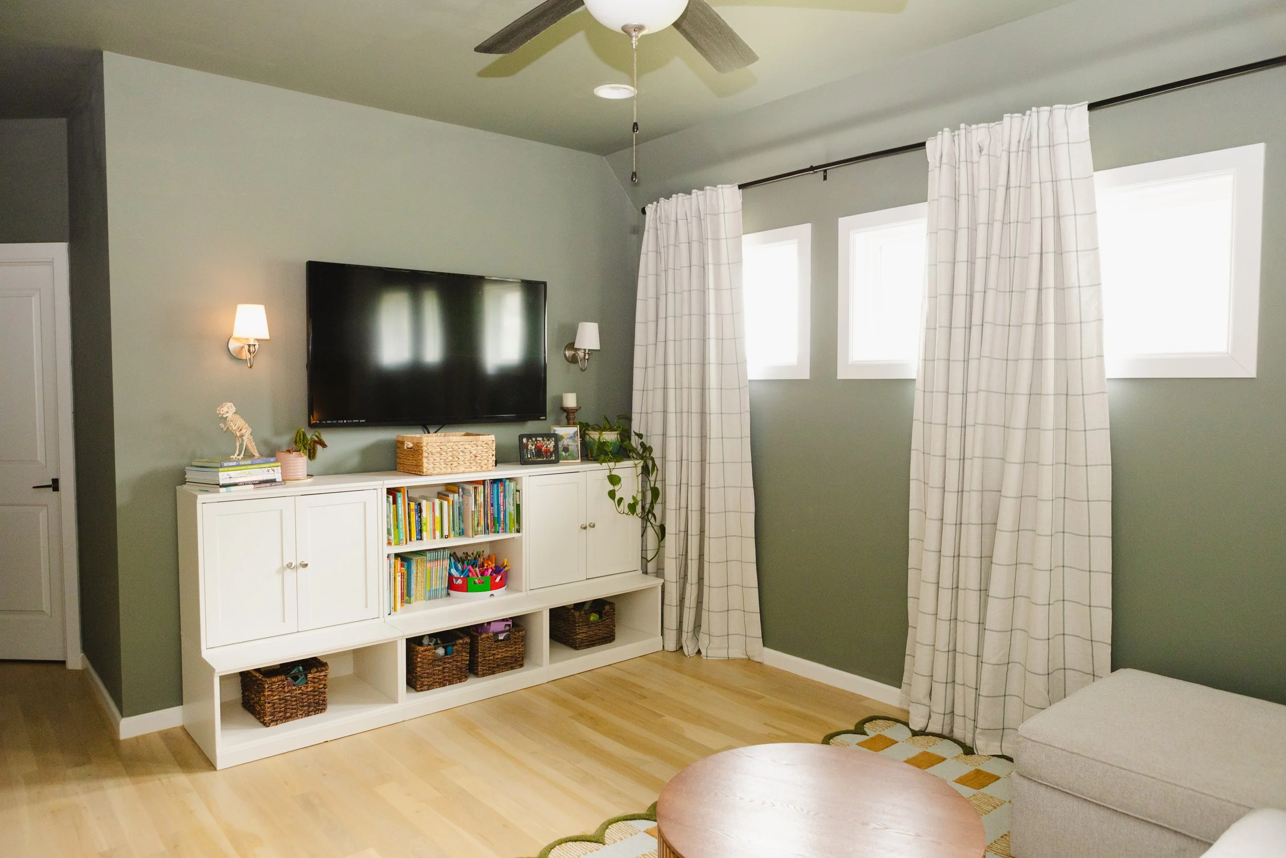 A living room area with a white bookshelf, a flat-screen TV mounted on a pale green wall, and white curtains covering large windows. There are two wall-mounted lamps, a small plant, framed photos, and a bibelot on top of the bookshelf. Part of a sitt
