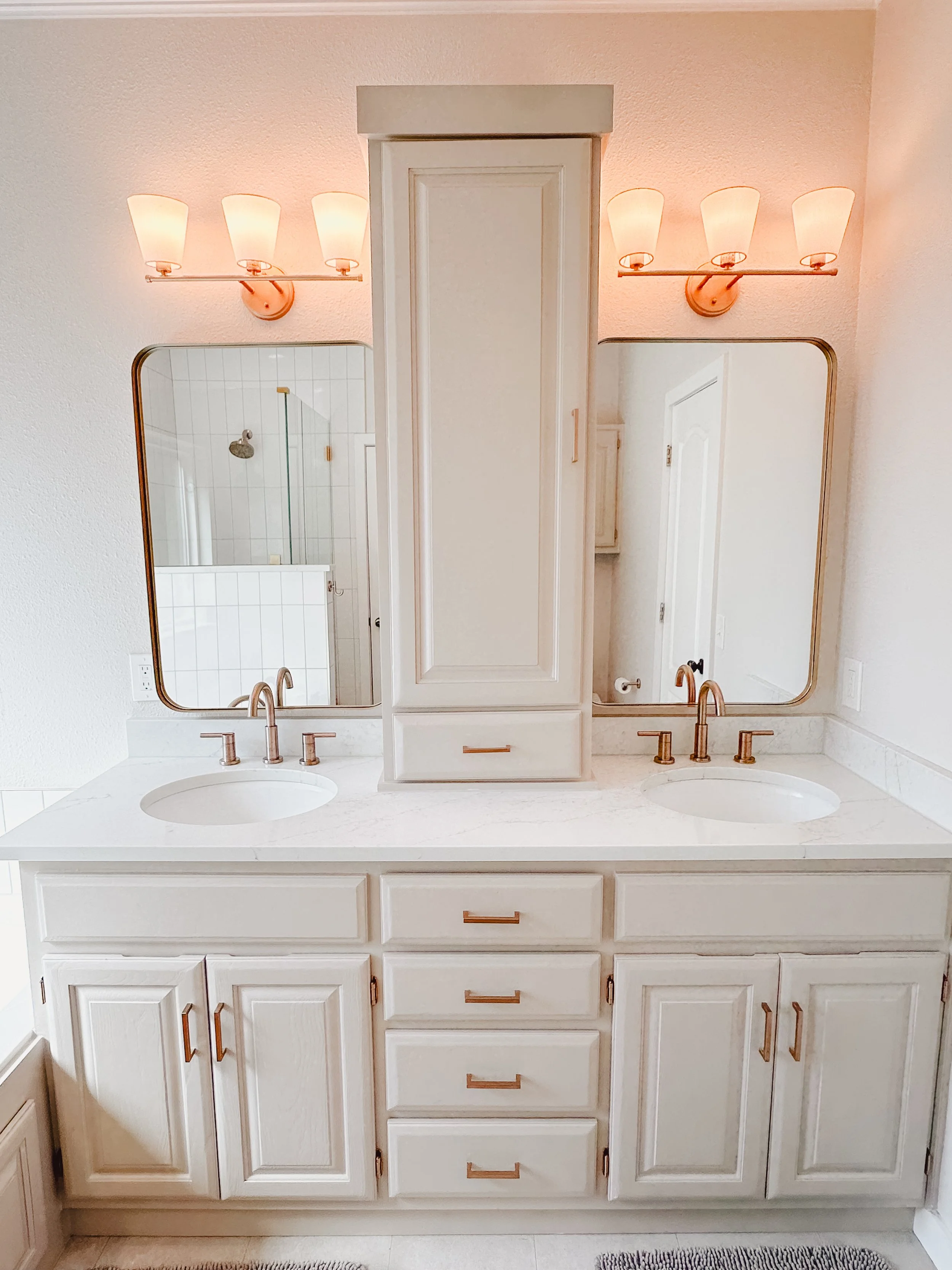 Double vanity bathroom with two sinks, white cabinets, large mirror, brass fixtures, and a central cabinet with drawers, in a neutral-colored room.