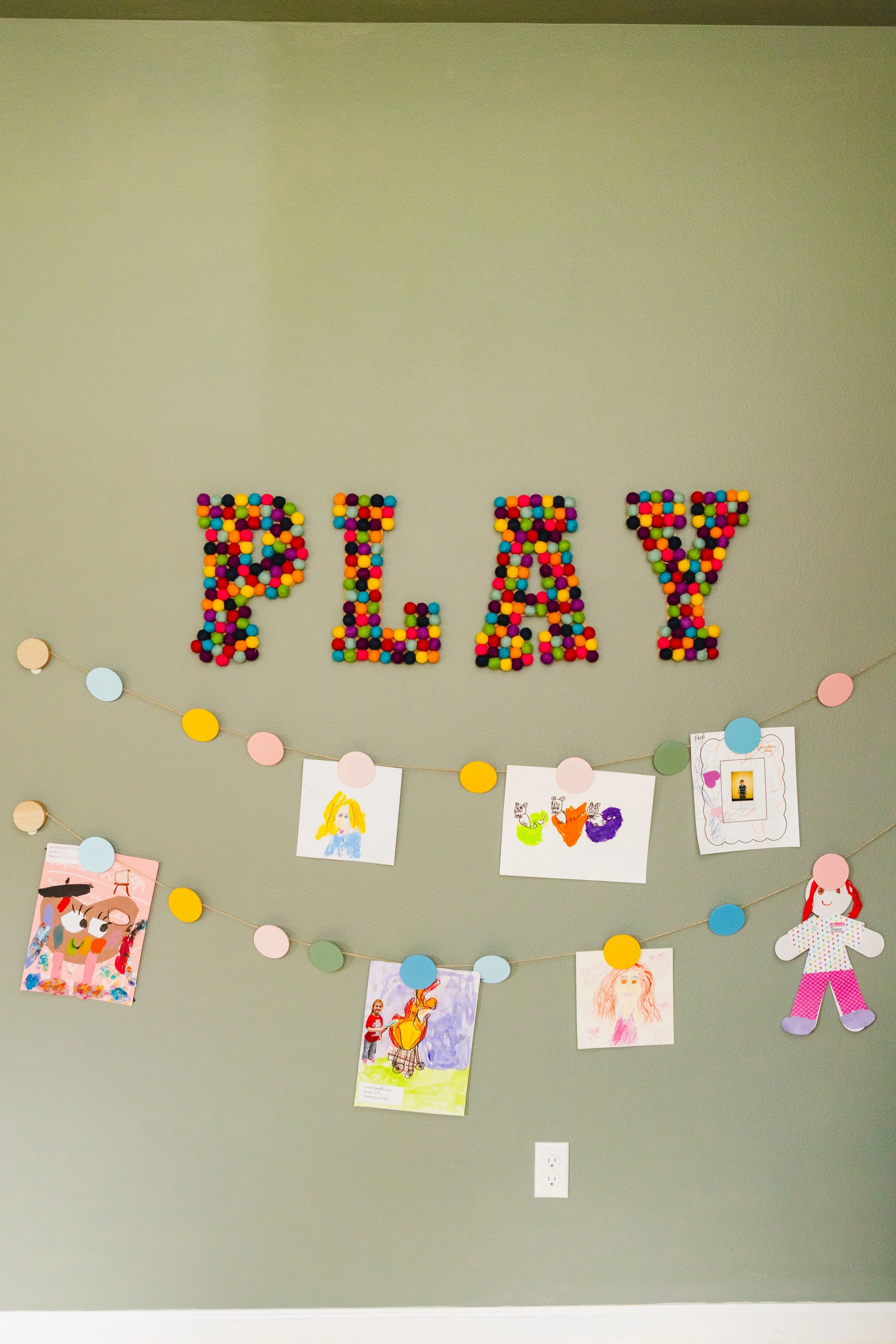 Wall decoration with the word 'PLAY' spelled out with colorful letter-shaped candies. Below, children's drawings and artwork are displayed on a string with paper circles.