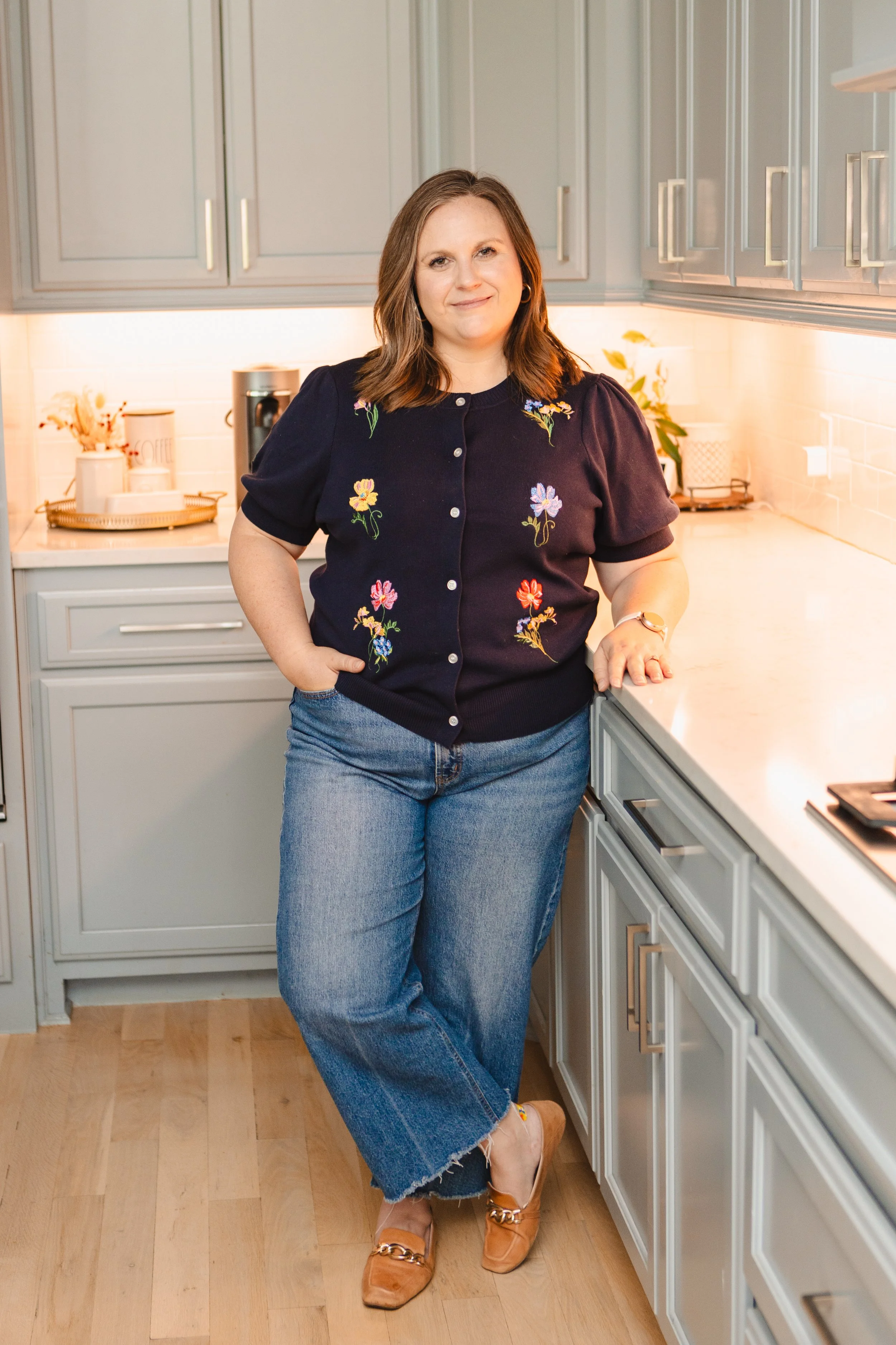 A woman standing in a kitchen with her arms resting on the countertop, wearing a navy sweater with floral embroidery, blue jeans, and tan shoes, smiling at the camera.