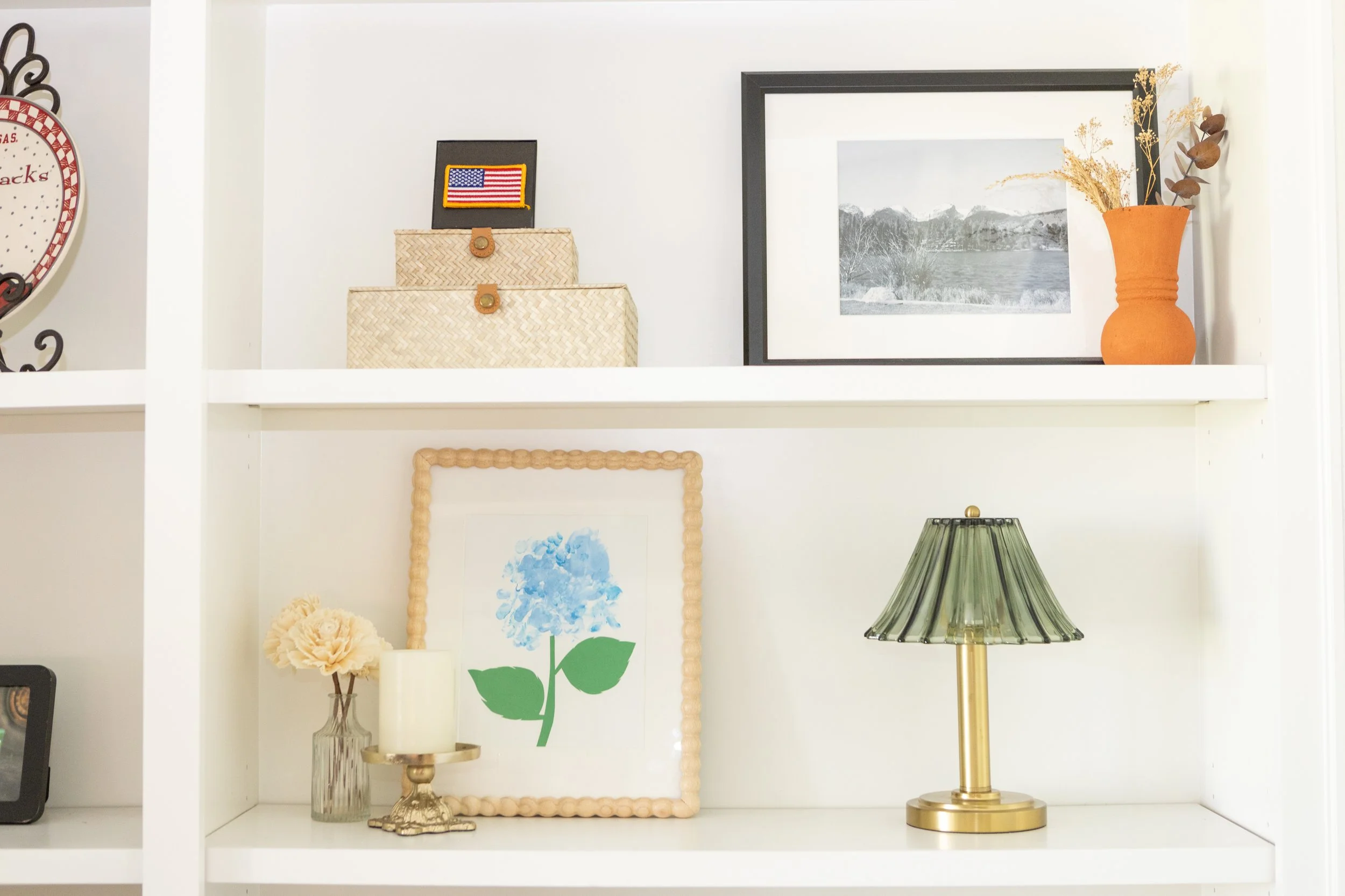 Decorative shelf with framed art, a vase with dried flowers, a gold lamp with a green shade, and a few small ornaments.