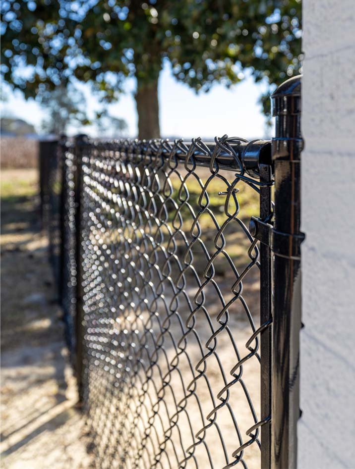 Close-up of a black chain-link fence attached to a concrete wall, with a blurred background of trees and sky.