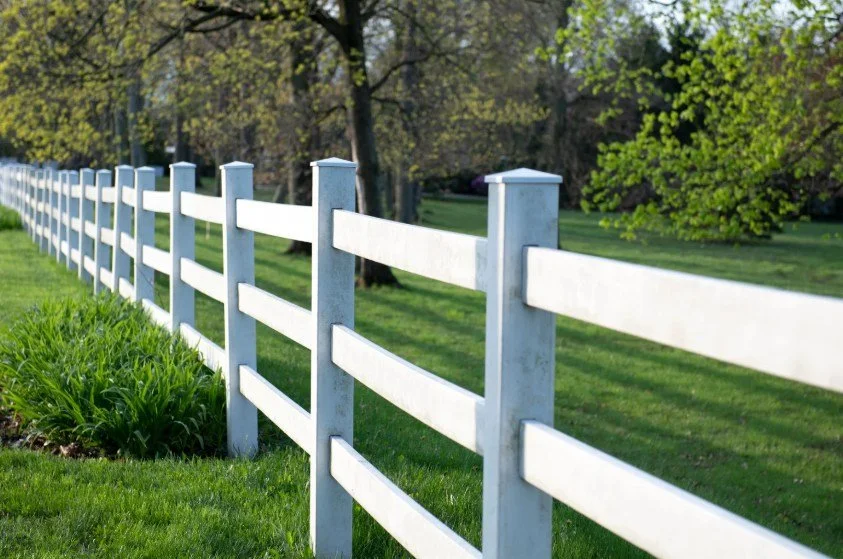 White PVC fence running through a green grassy field with trees in the background.