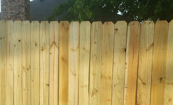 A wooden privacy fence made of light-colored vertical planks, with a tree and part of a house visible in the background.
