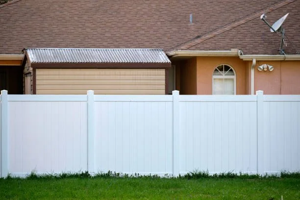 View of the backyard PVC fence with a house in the background, featuring a small garage with a metal roof, a satellite dish, and an arched window.