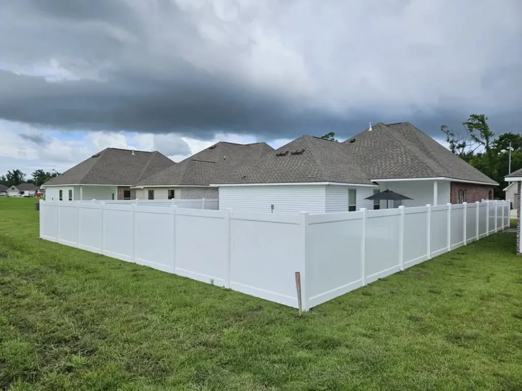 Backyard with white vinyl privacy fence surrounding a house with gray shingle roof, on a cloudy day.