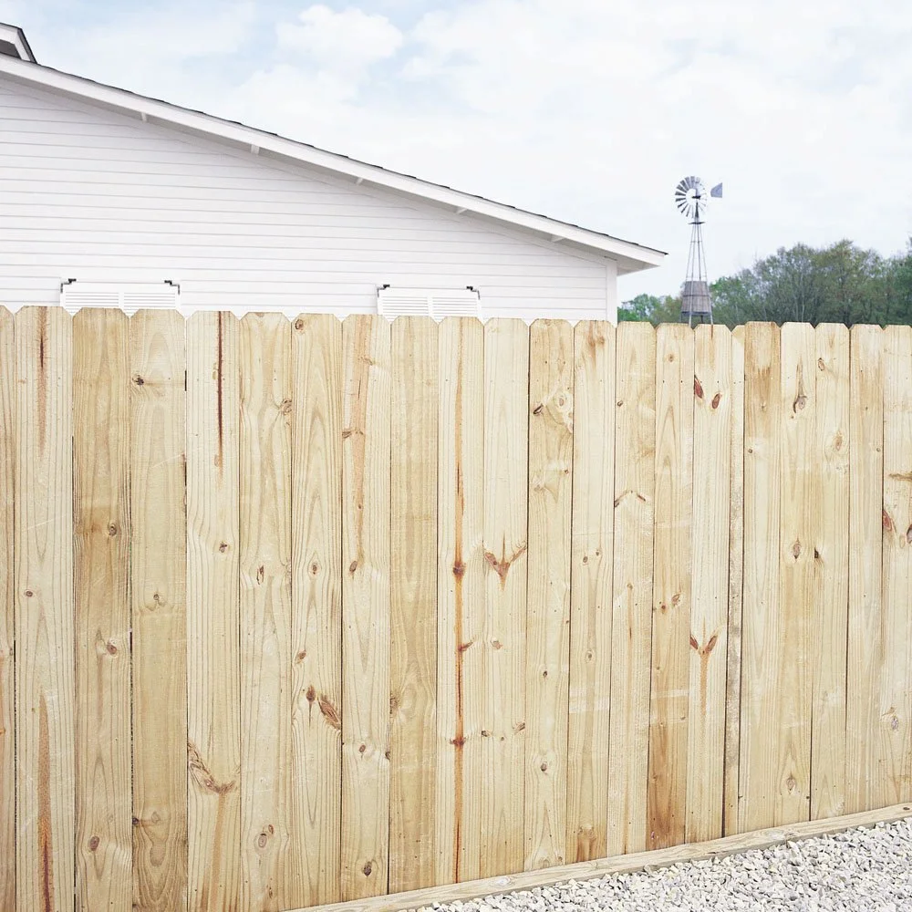 A new wooden privacy fence in front of a white house with a sloped roof and a windmill in the background on a cloudy day.