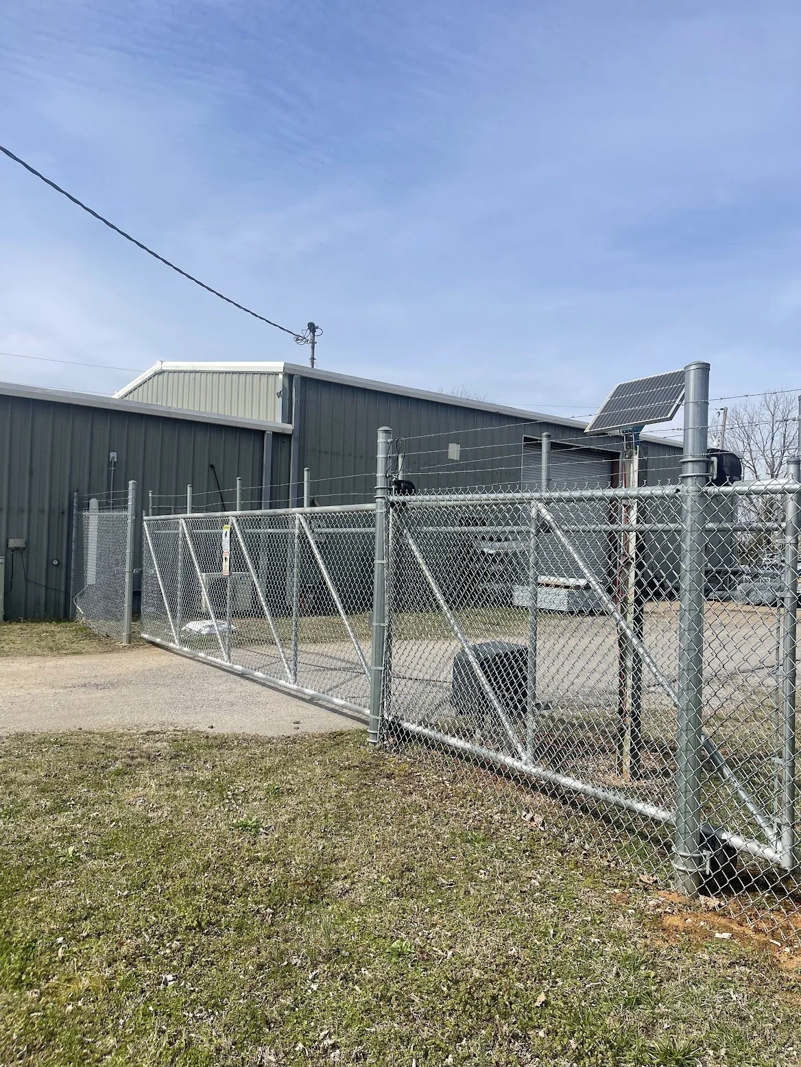 Industrial building with a chain-link fence, solar panel, and cantilever gate outside under a blue sky.