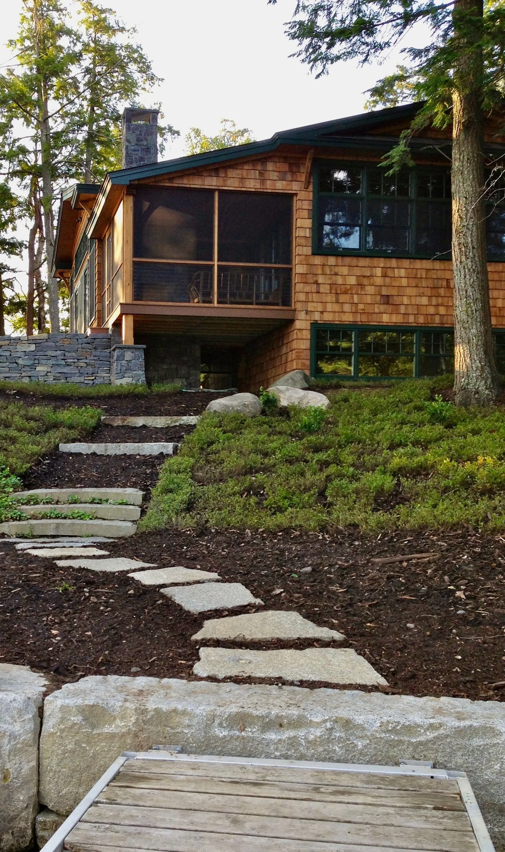 A stone pathway leads up a hill to a wooden house with large windows and a screened porch, surrounded by trees.