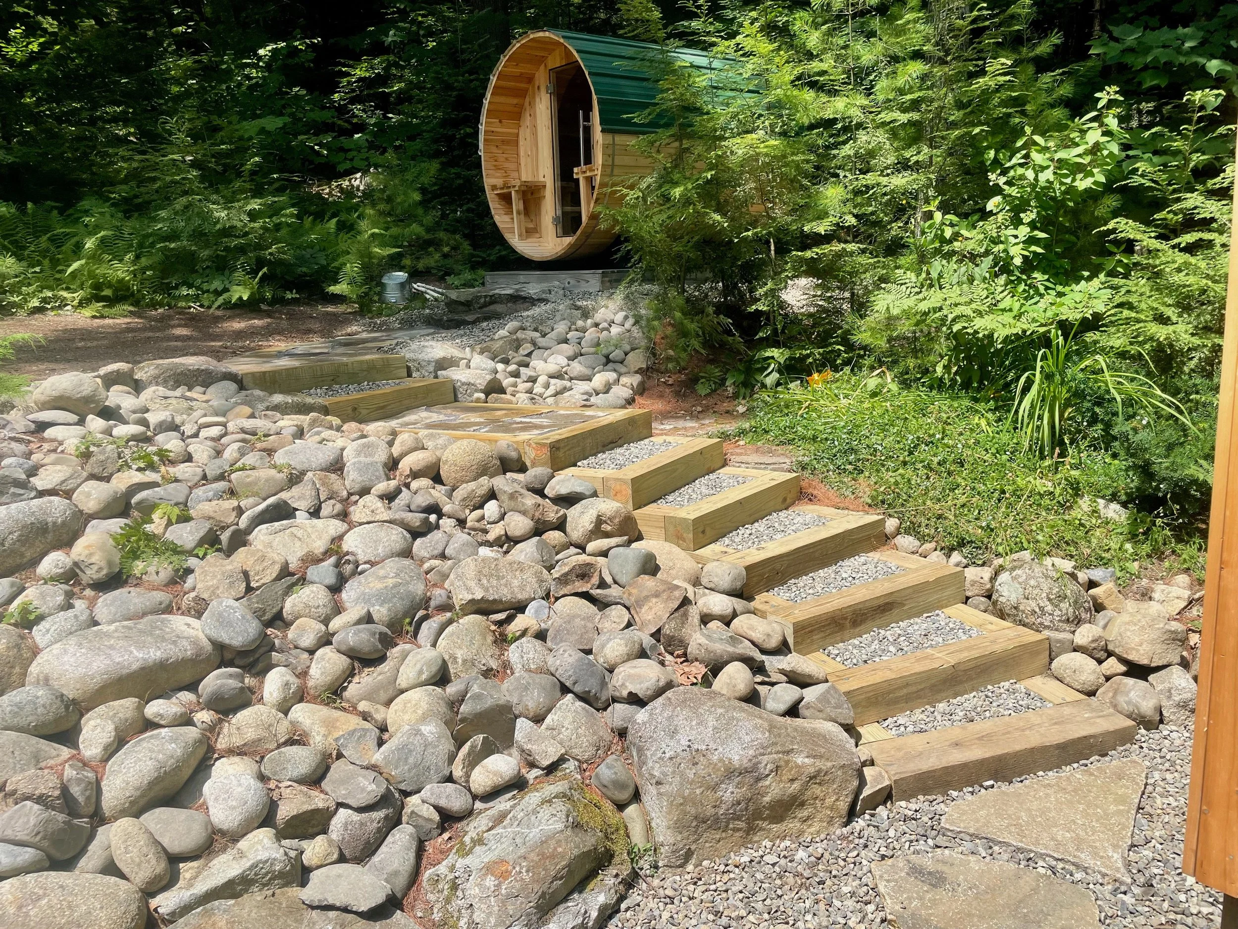 A small wooden barrel-shaped sauna with a green metal roof, situated on a wooden platform surrounded by lush green foliage and a pathway made of stones and wooden steps.