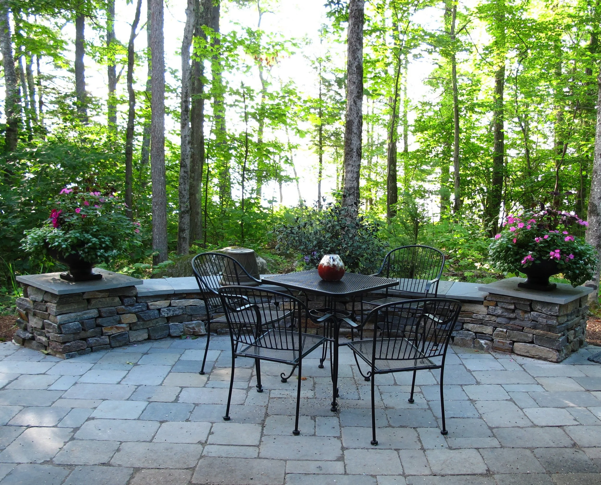 A backyard patio with black metal table and chairs, a stone brick wall, potted plants with pink flowers, and a lush green forested area in the background.
