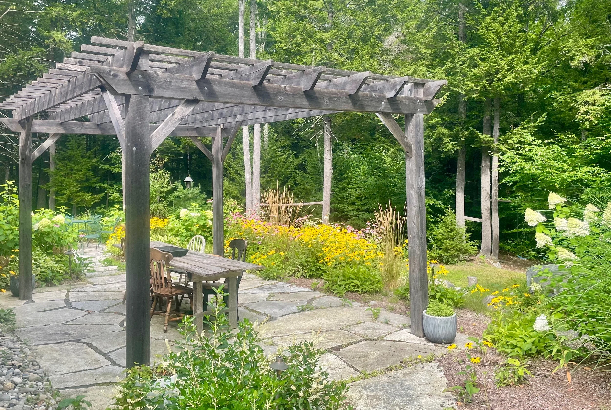 A wooden pergola over a stone patio in a lush garden with colorful flowers and trees.