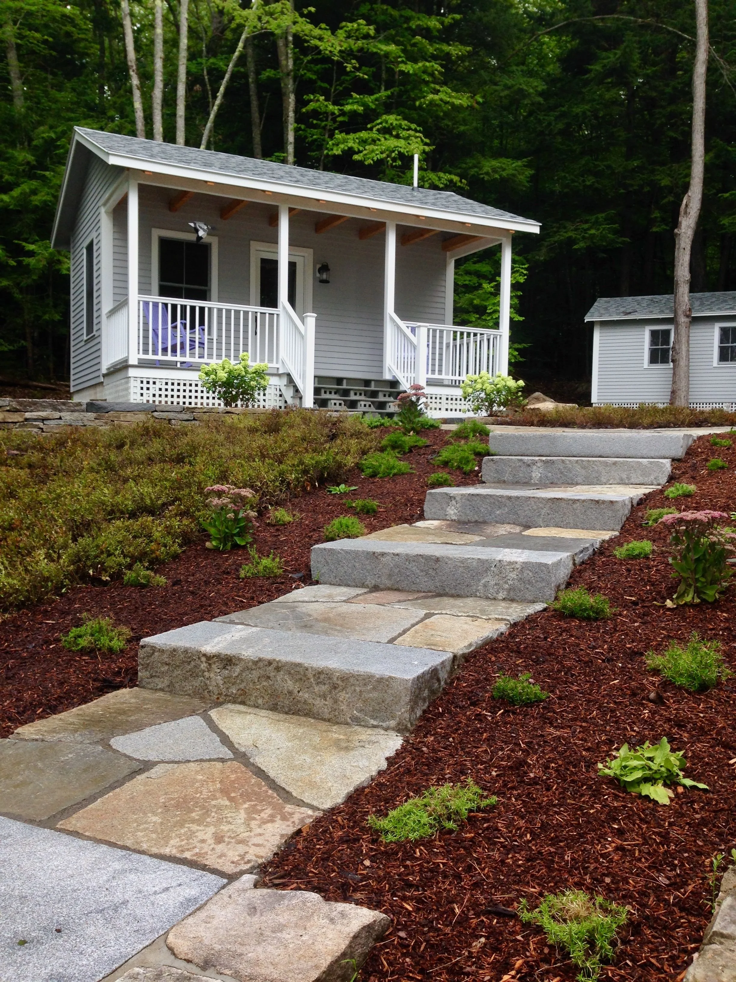A small white house with a porch and steps, surrounded by a landscaped garden with mulch, plants, and a pathway made of stone steps, set against a backdrop of green trees and forest.