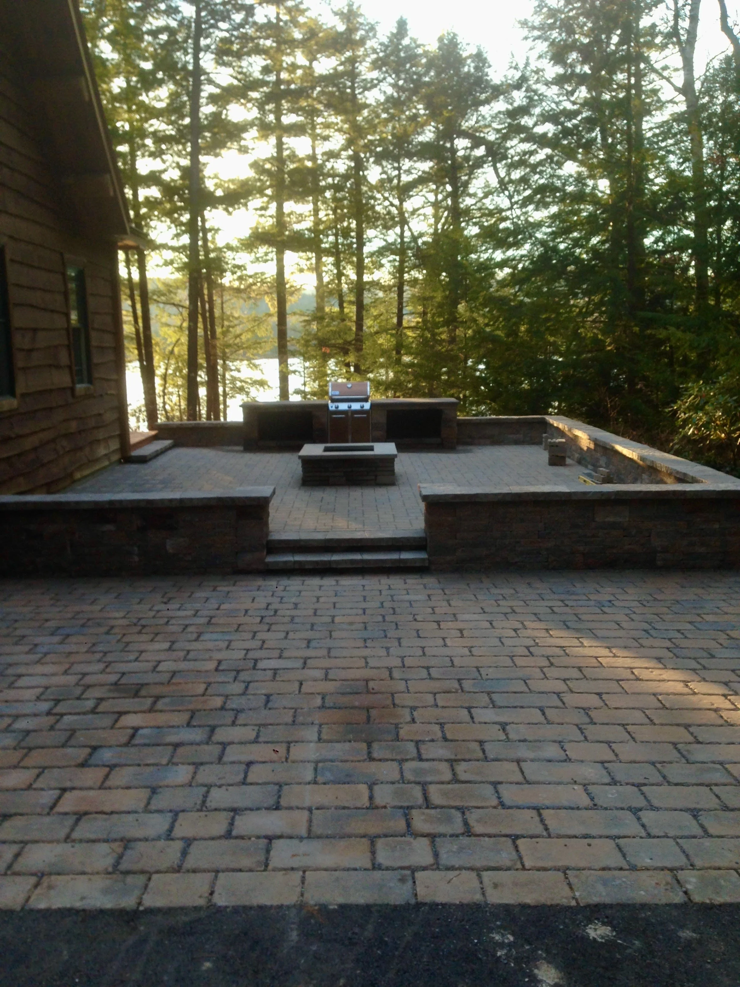 Backyard patio with brick flooring, built-in brick fire pit, surrounded by benches, and a view of a wooded area with tall trees in the background.