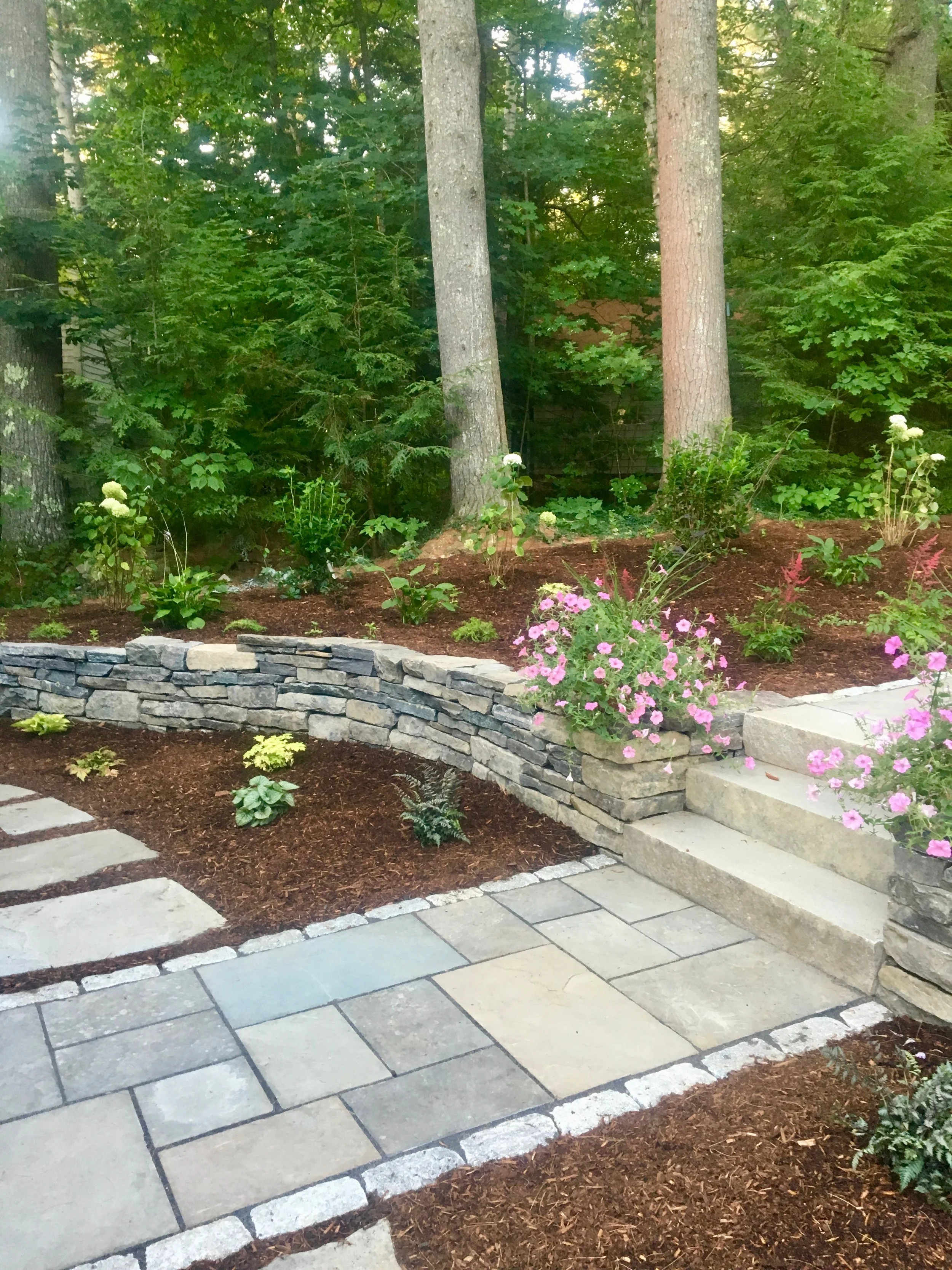 A landscaped garden with a tiered flower bed, stone retaining wall, and a paved pathway.