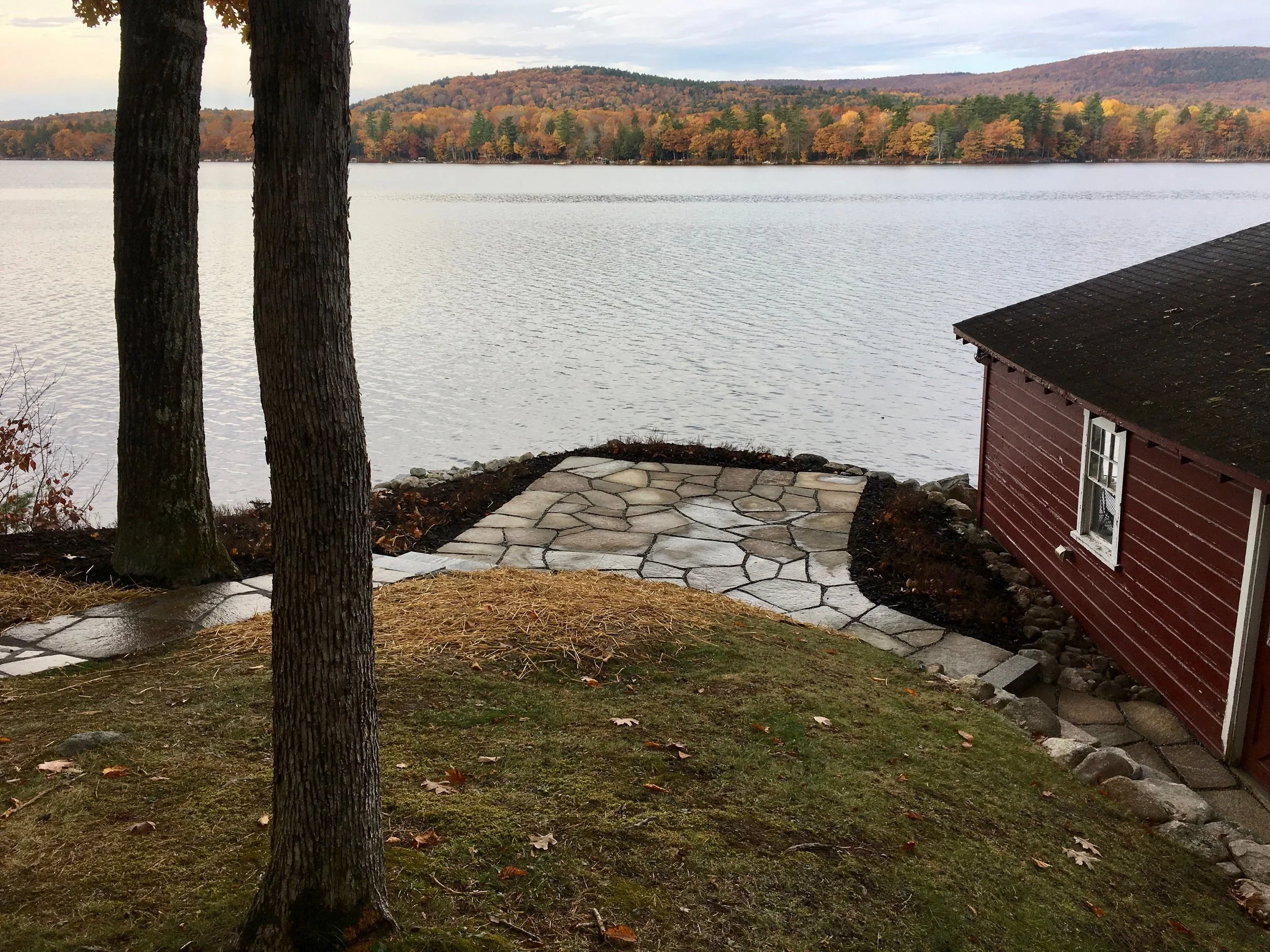 View of a lakeside in autumn with trees displaying fall colors, a stone pathway, and part of a red house with a dark roof.