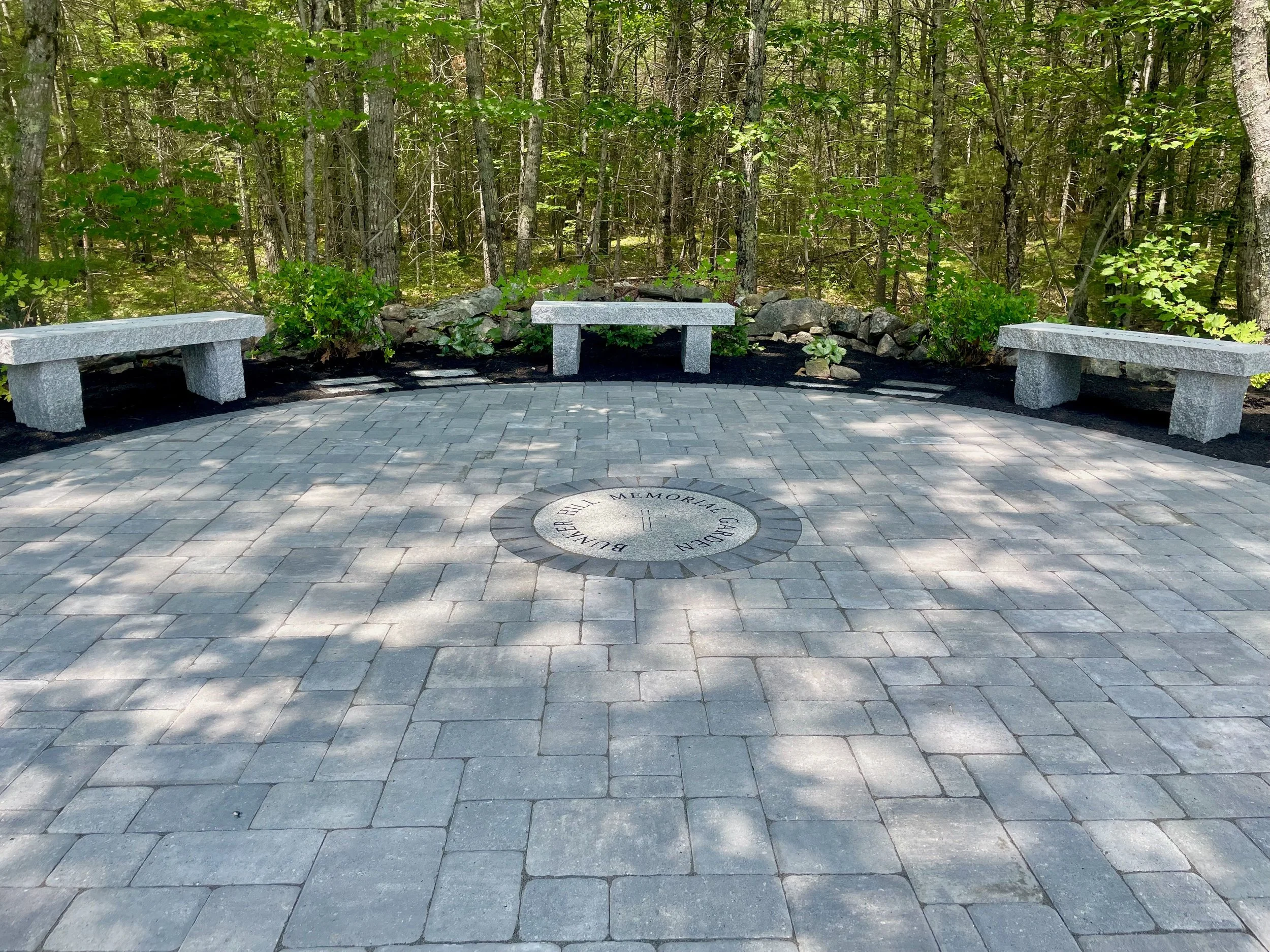 A memorial plaque embedded in a paved area with three stone benches around it, located in a forest with tall trees and green foliage.