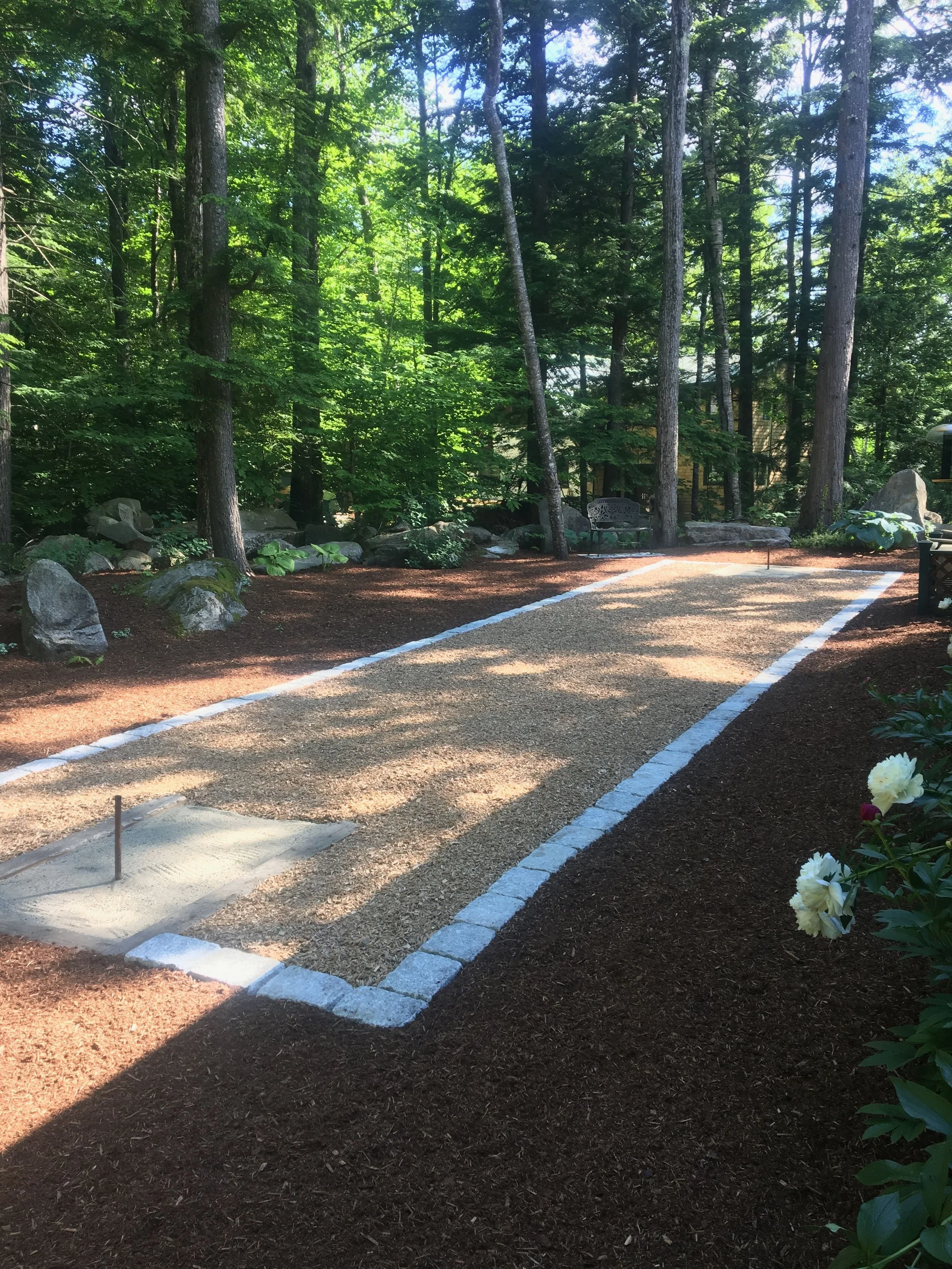 A miniature golf course with a sand trap in the foreground, brown mulch on the ground, surrounded by large rocks and green plants, with a wooded area with tall trees in the background.