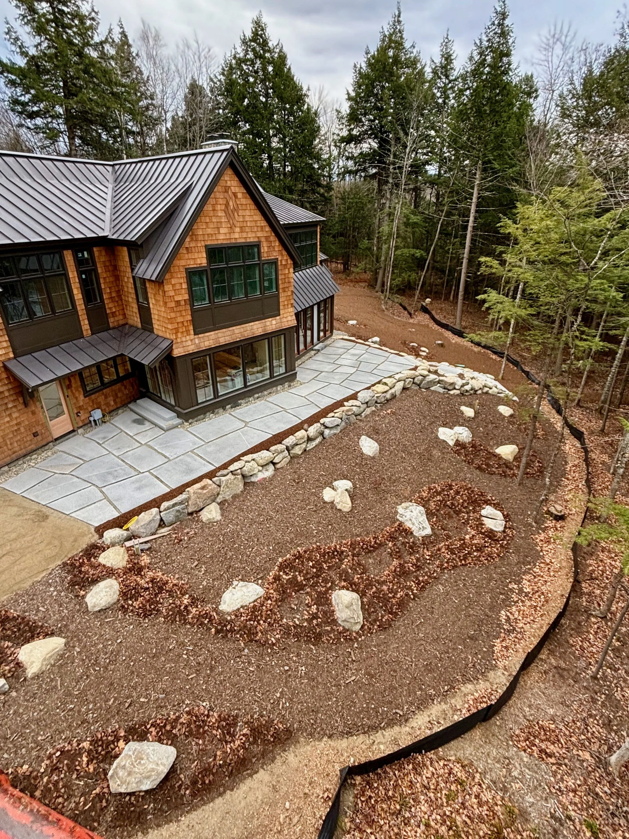 A house with a metal roof and large windows, surrounded by a newly landscaped yard with rocks, mulch, and a designated garden area, with a forest of tall trees in the background.