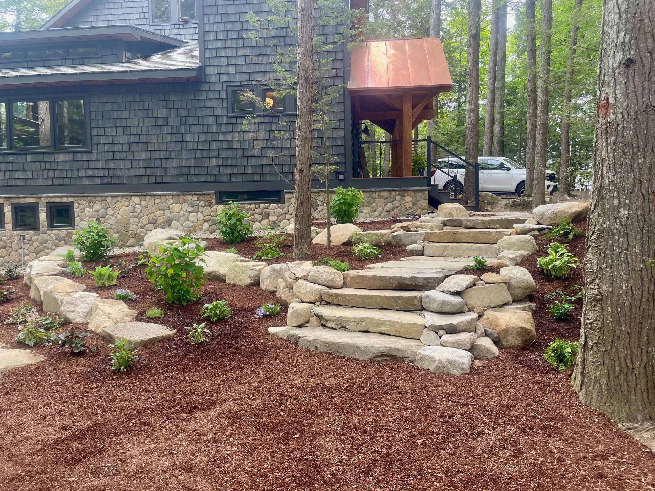 A house with black shingles, stone foundation, large windows, and a wooden porch with stairs leading up to it, surrounded by trees and a landscaped yard with small bushes and rocks.