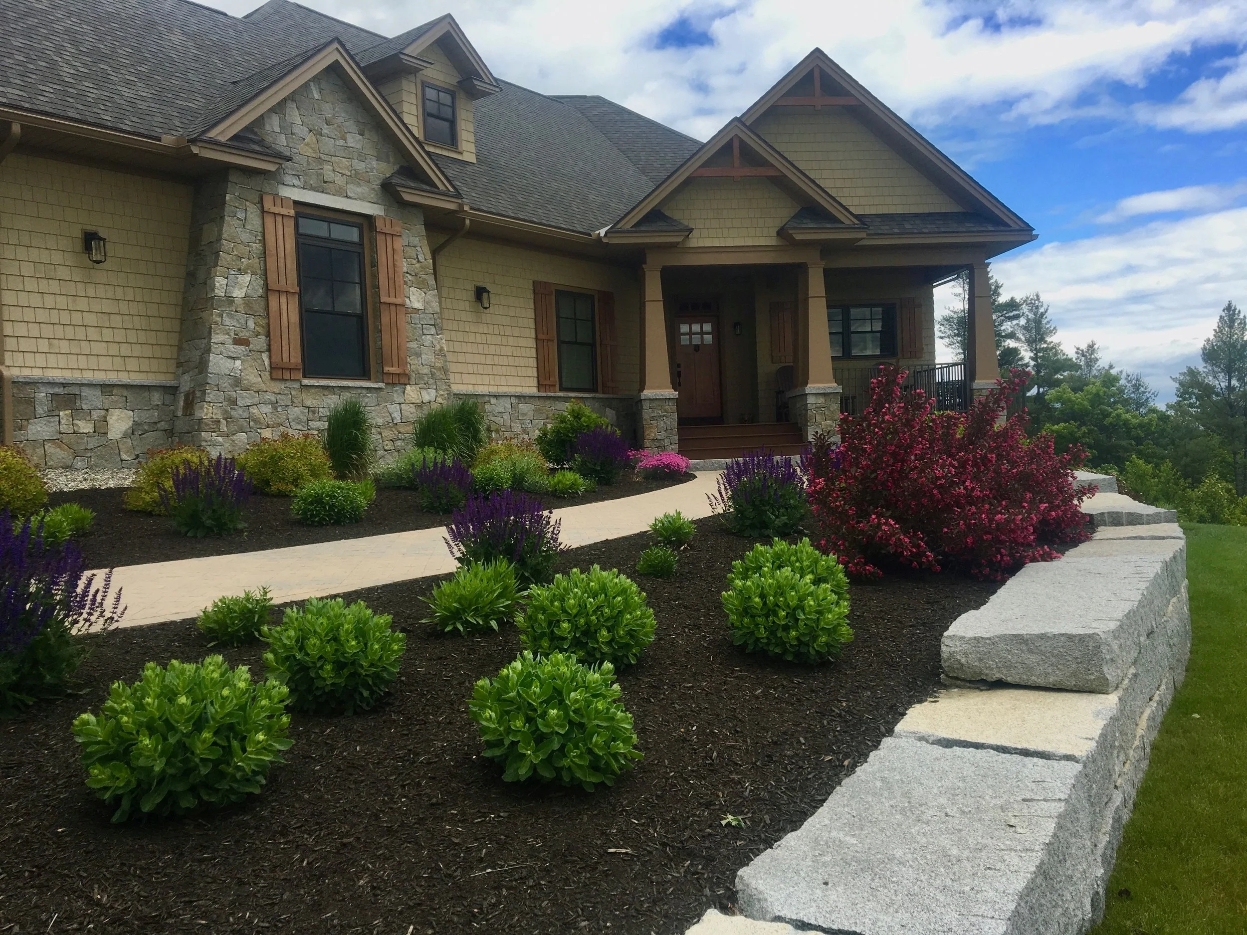 A large suburban house with beige siding, stone accents, and a steep gabled roof. The front yard features a landscaped garden with green bushes, purple flowers, and a curved walkway to the entrance, with tall trees in the background under a partly cl