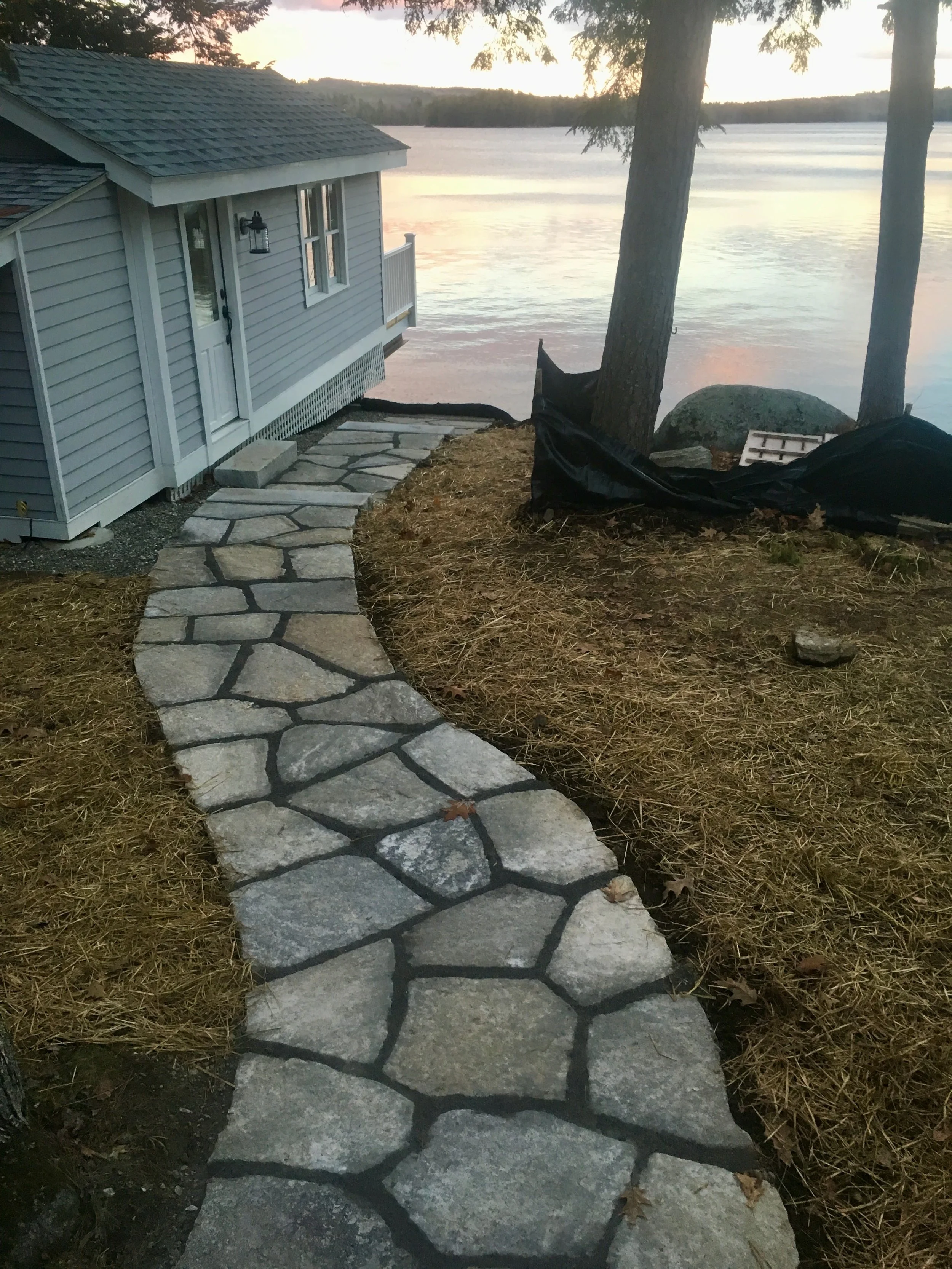 A stone pathway leading away from a house towards a body of water in a scenic outdoor setting during twilight, with trees, a large rock, and a black tarp or fabric on the ground.