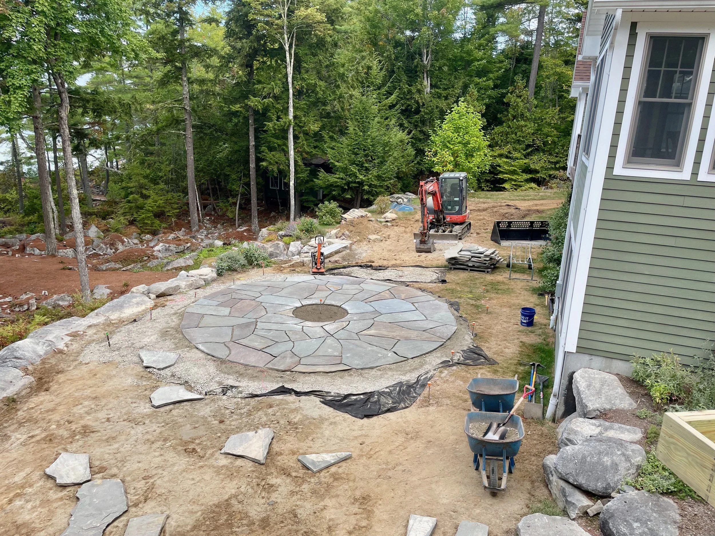 A backyard under landscaping renovation with a stone patio, a mini excavator, and construction supplies near a house with green siding, surrounded by tall trees.