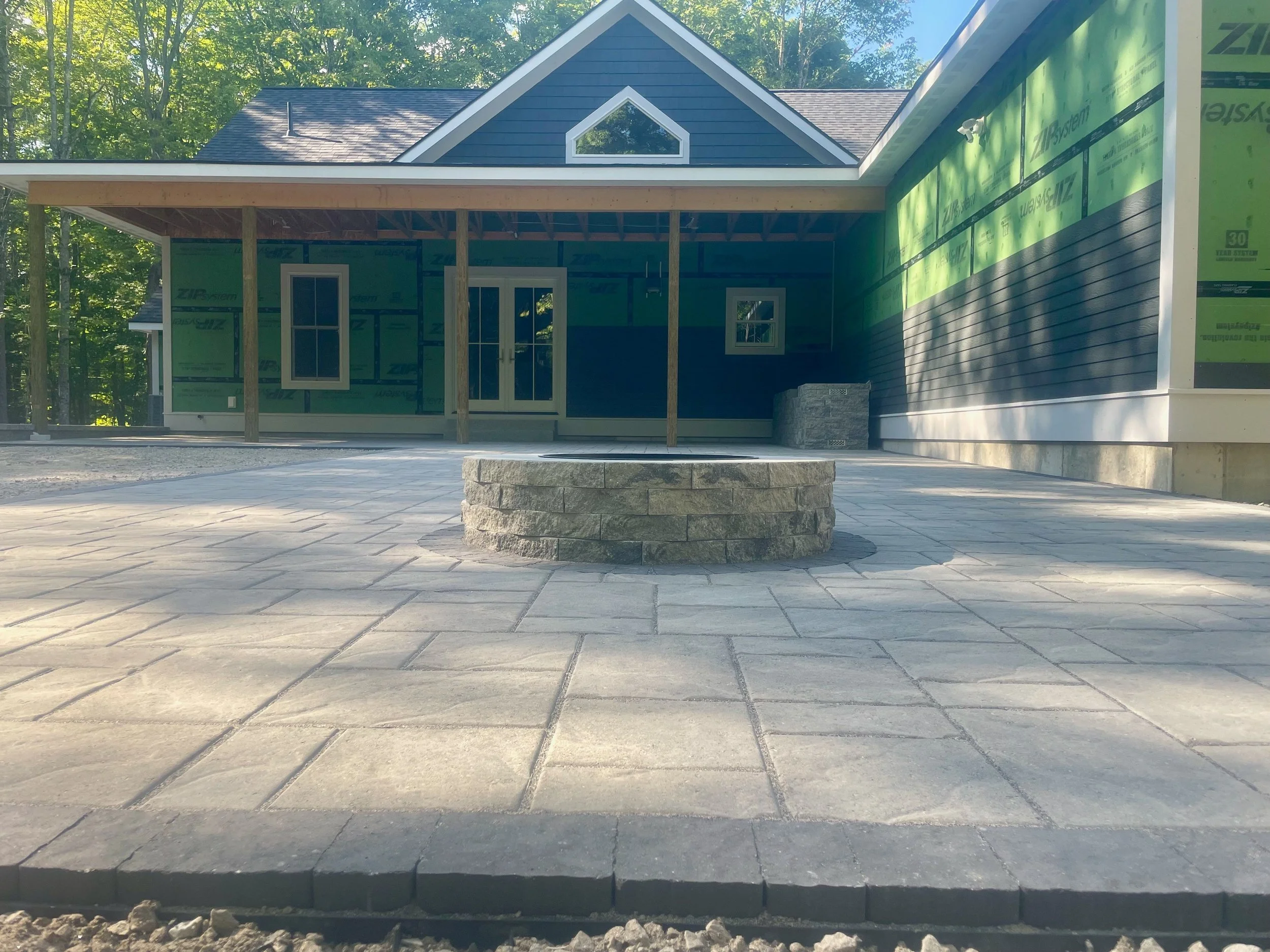 Exterior of a house under construction, with a blue facade, covered porch, and two small windows. The porch is supported by wooden posts, and there is a stack of stone near the house. The area is paved with stone tiles, and trees are visible in the b