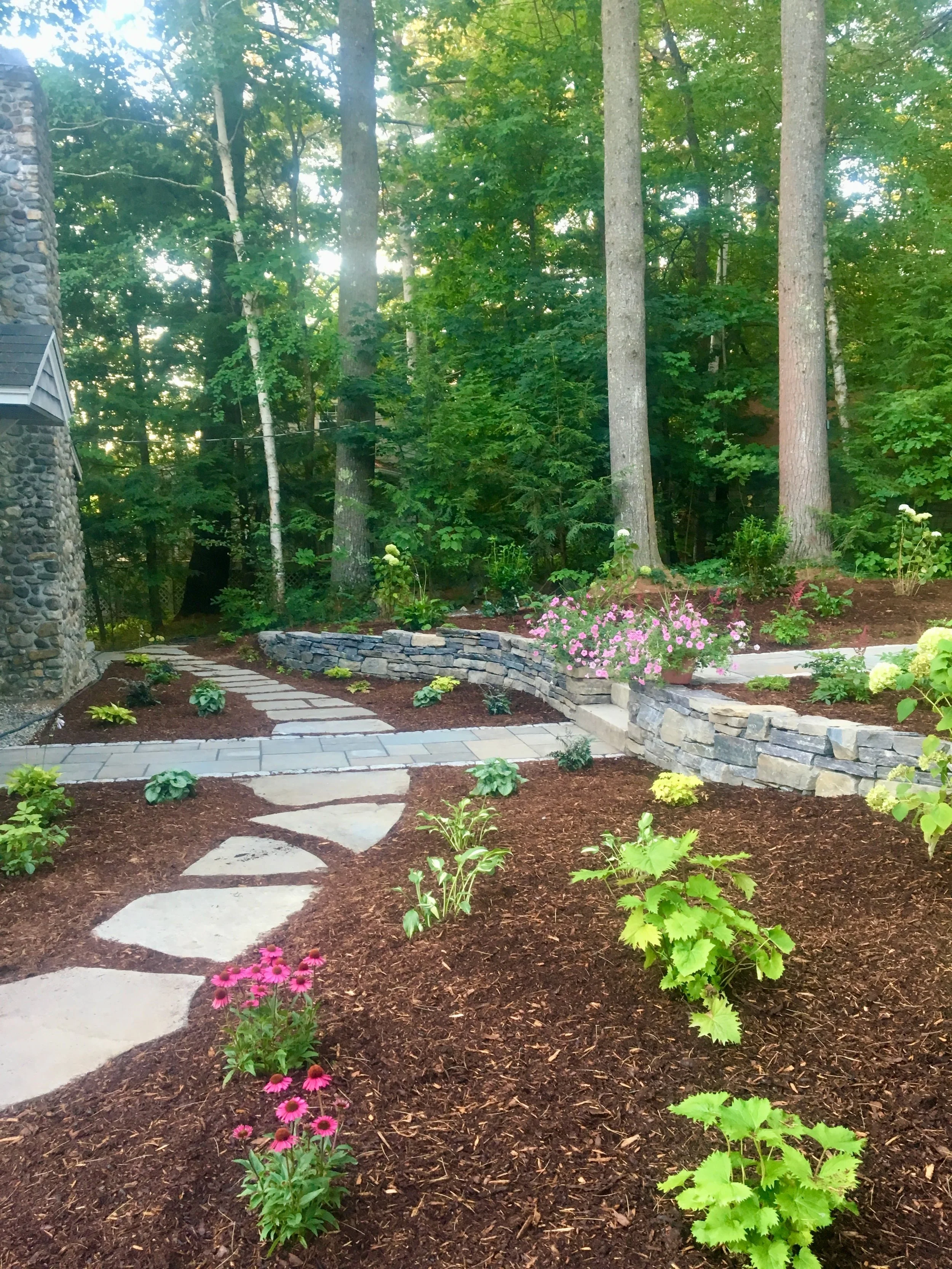 A landscaped garden with a stone pathway, colorful flowers, a stone wall, and tall trees in the background.