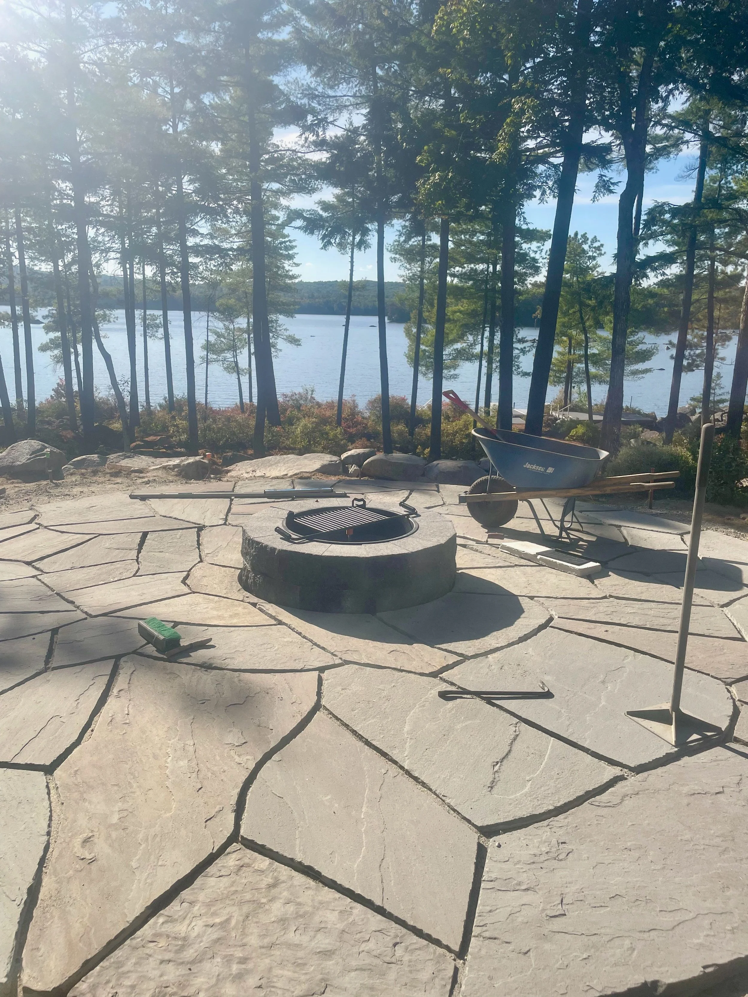 View of a lakeside scene with trees in the background, a stone patio with fire pit, wheelbarrow, and various tools in the foreground.