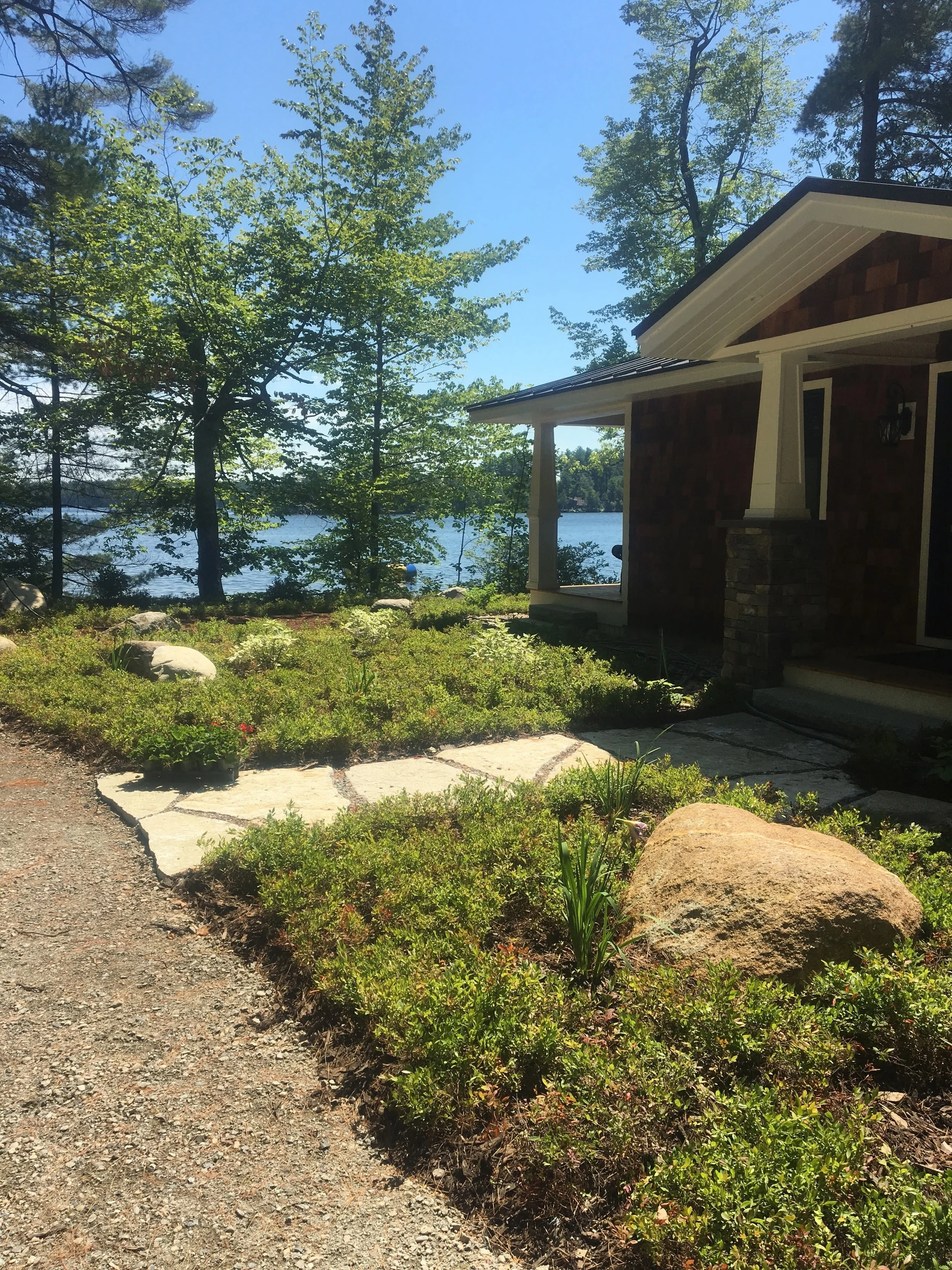 A house near a lake with trees and bushes in the foreground, clear blue sky above.
