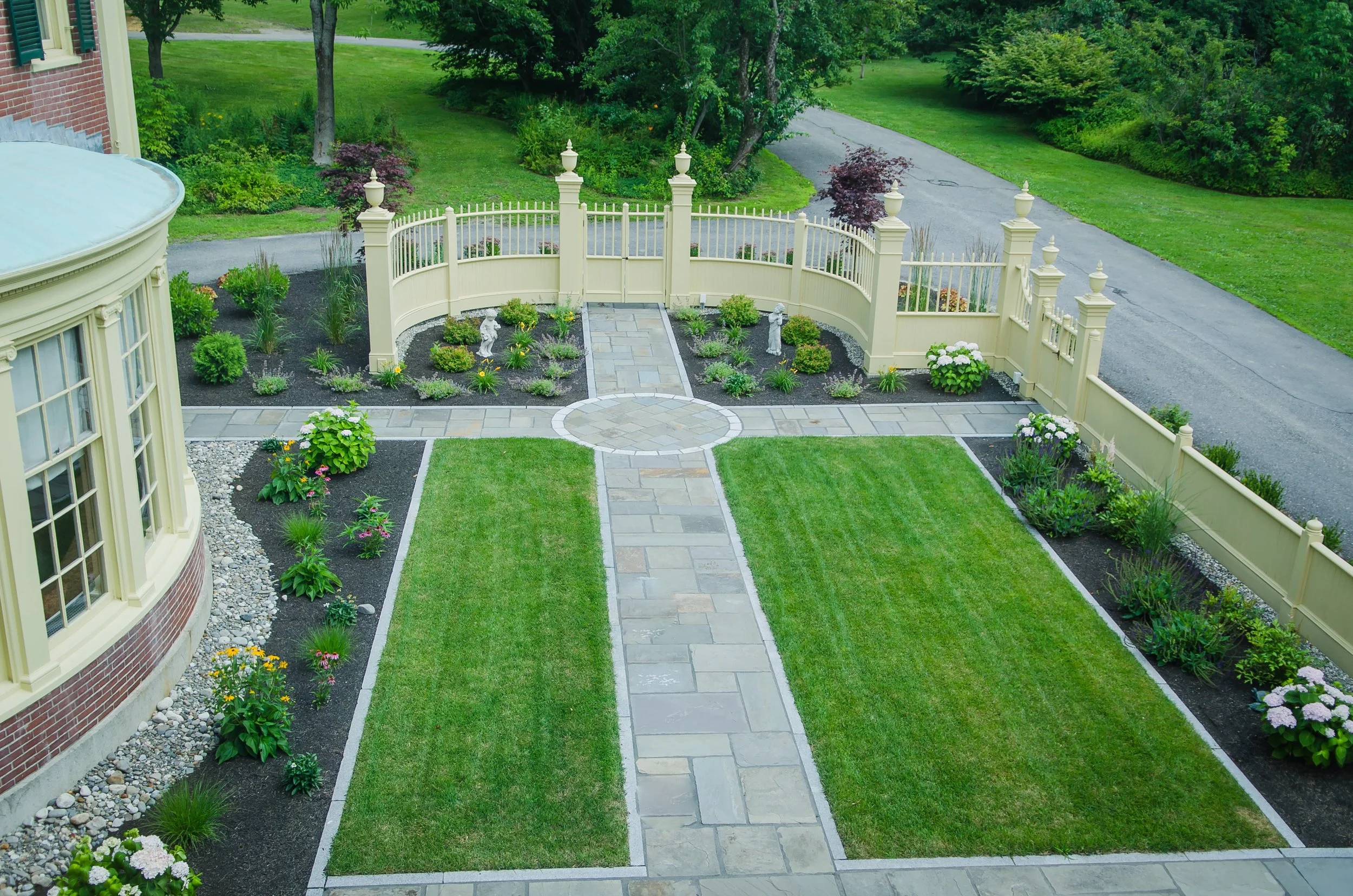 A front yard with a stone pathway leading to a white picket fence with decorative posts and urn finials. The yard has landscaped flower beds with various plants and flowering shrubs, and a green lawn. In the background, there are trees and a road.