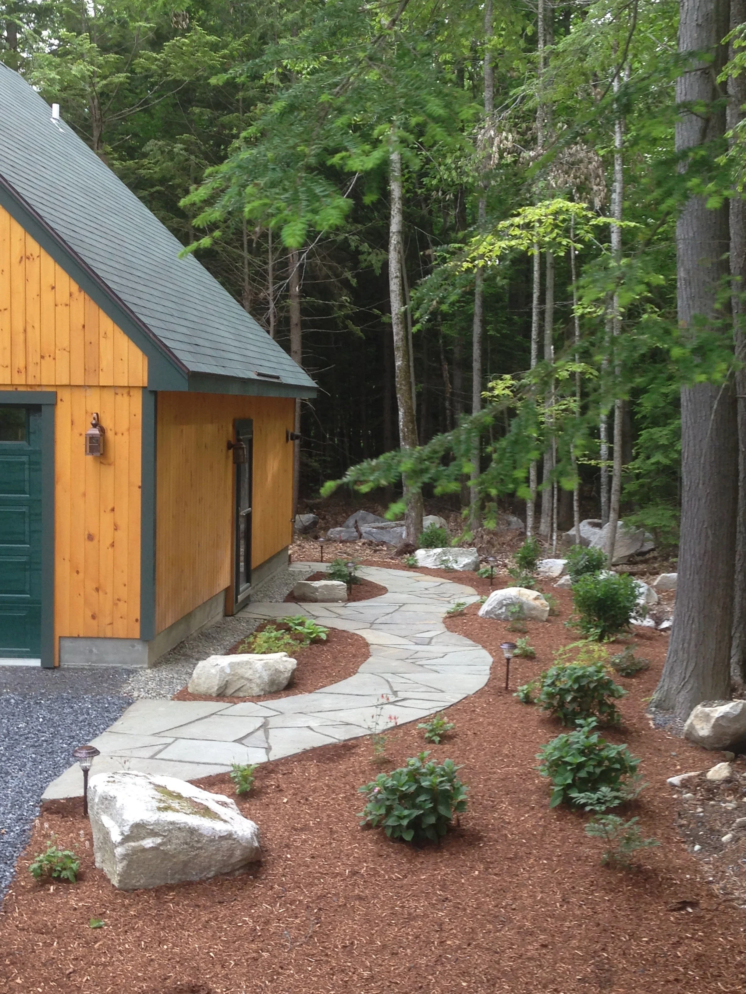A landscaped backyard with a winding stone pathway, small bushes, and large rocks, bordered by reddish mulch, leading to a wooded forest area, with a wooden house visible on the left side.