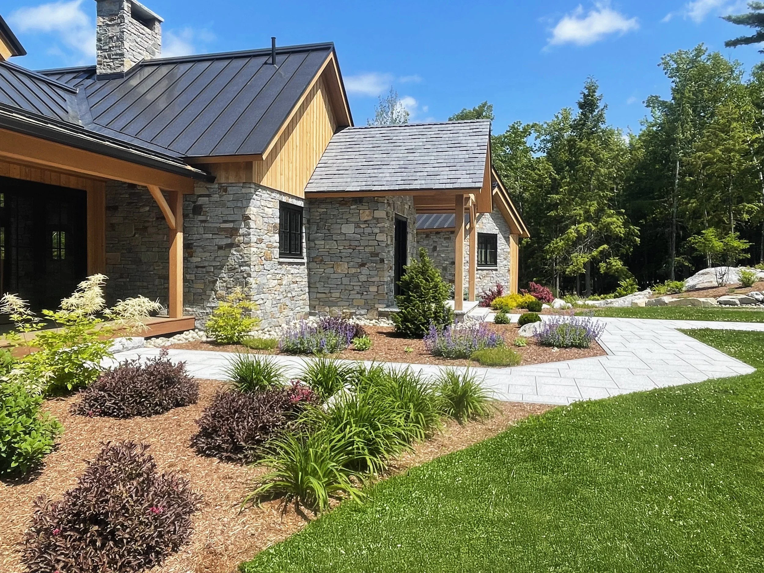 A modern house with a combination of stone and wood exterior, surrounded by landscaped garden with flowering plants and a stone pathway, under a clear blue sky.