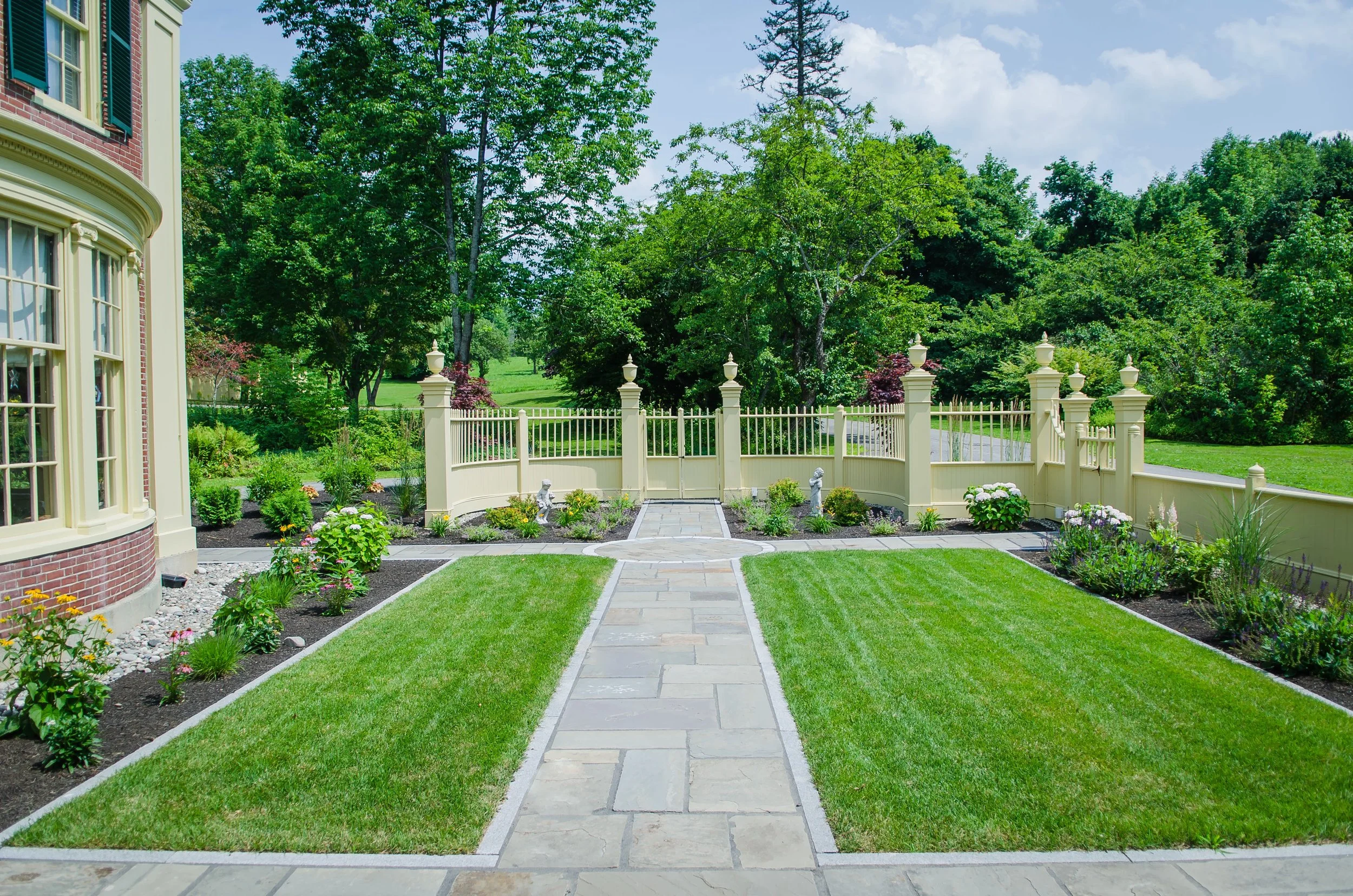 A landscaped backyard with a stone pathway leading to a white fence with decorative posts, surrounded by green grass, flower beds, and trees under a partly cloudy sky.